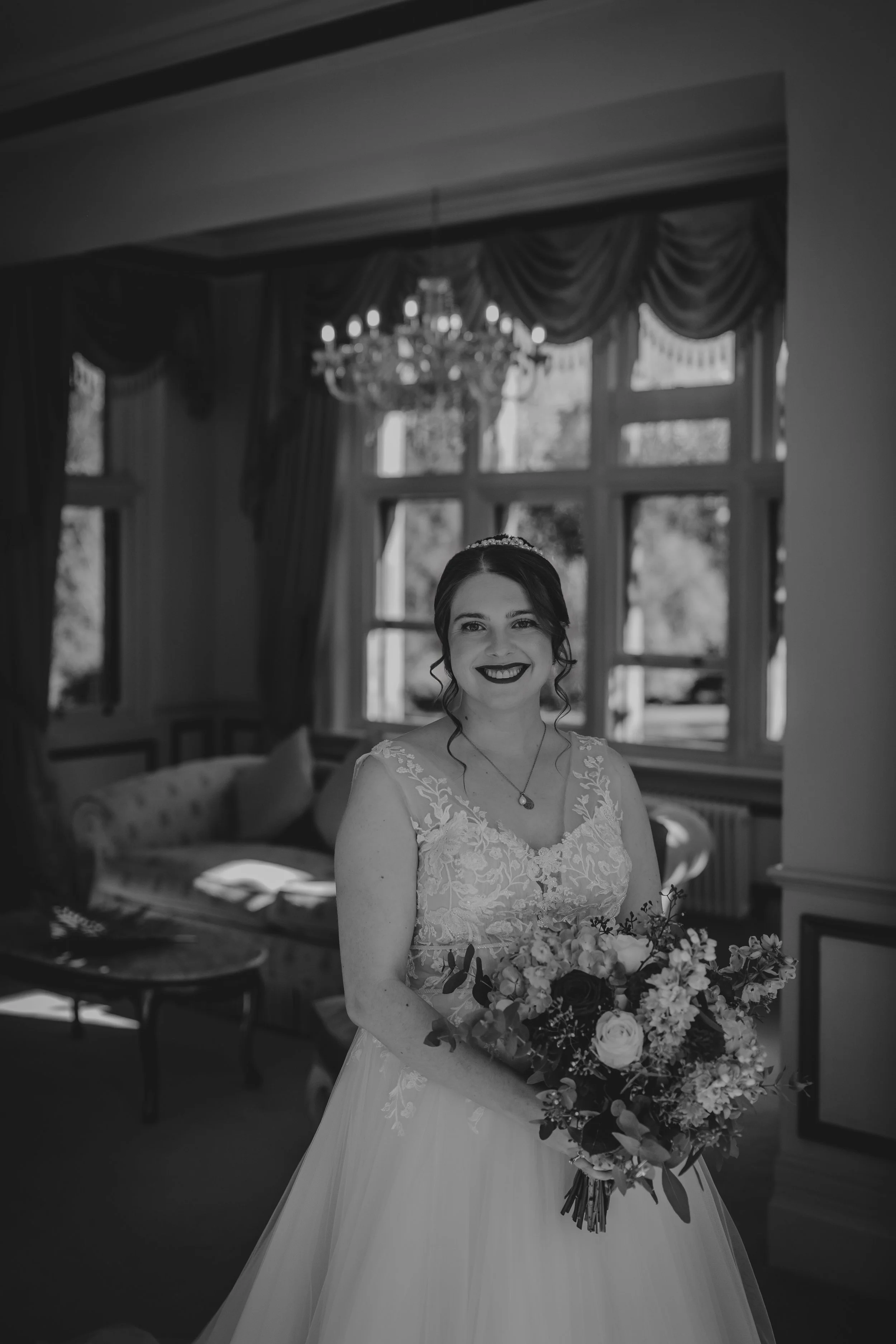 Black and white photo of a smiling bride holding a bouquet of flowers in a room with large windows, draped curtains, and a chandelier.