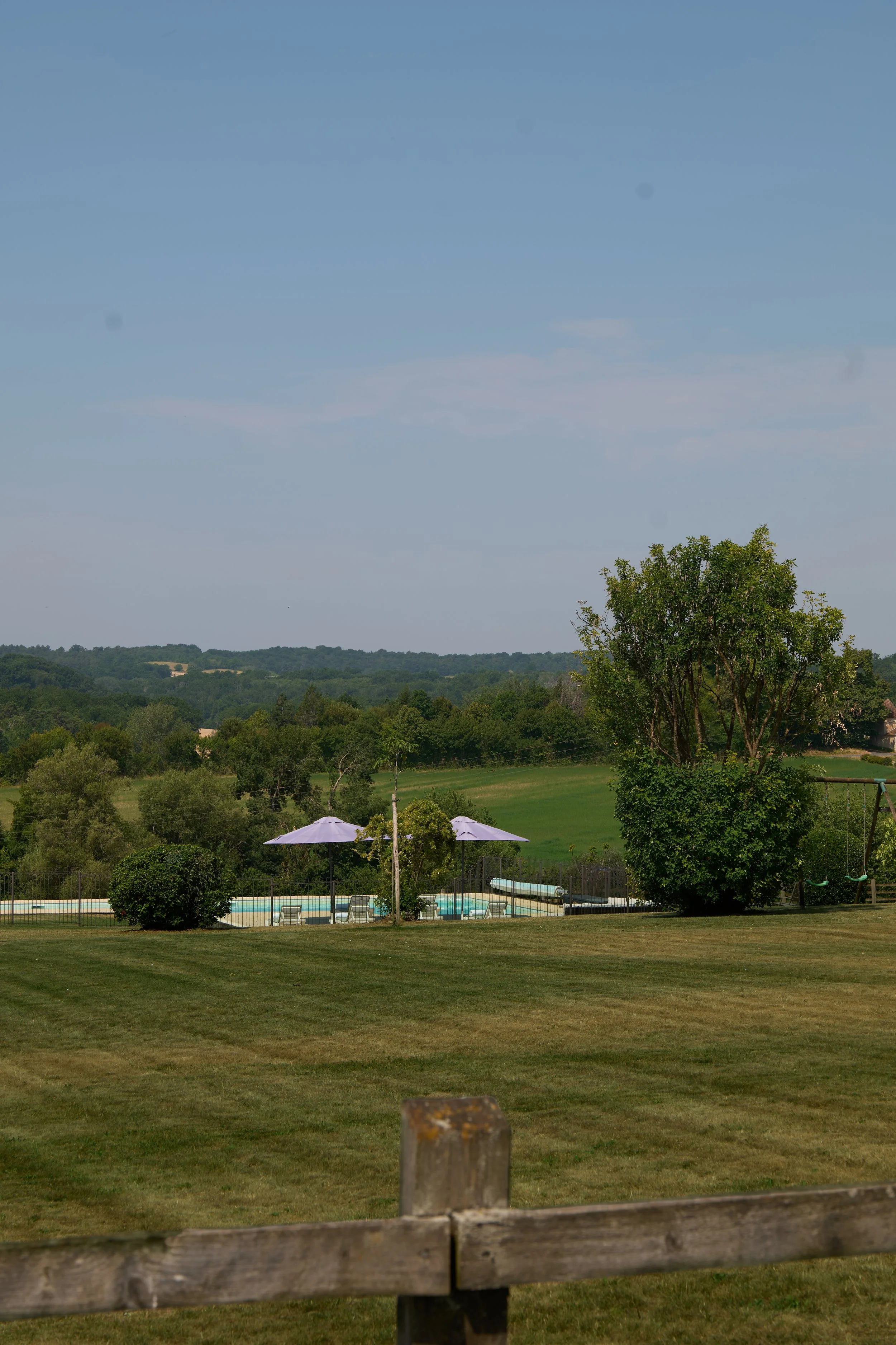 A scenic outdoor landscape showing a pool area with umbrellas and chairs, surrounded by grassy fields, trees, and distant hills under a blue sky with some clouds.