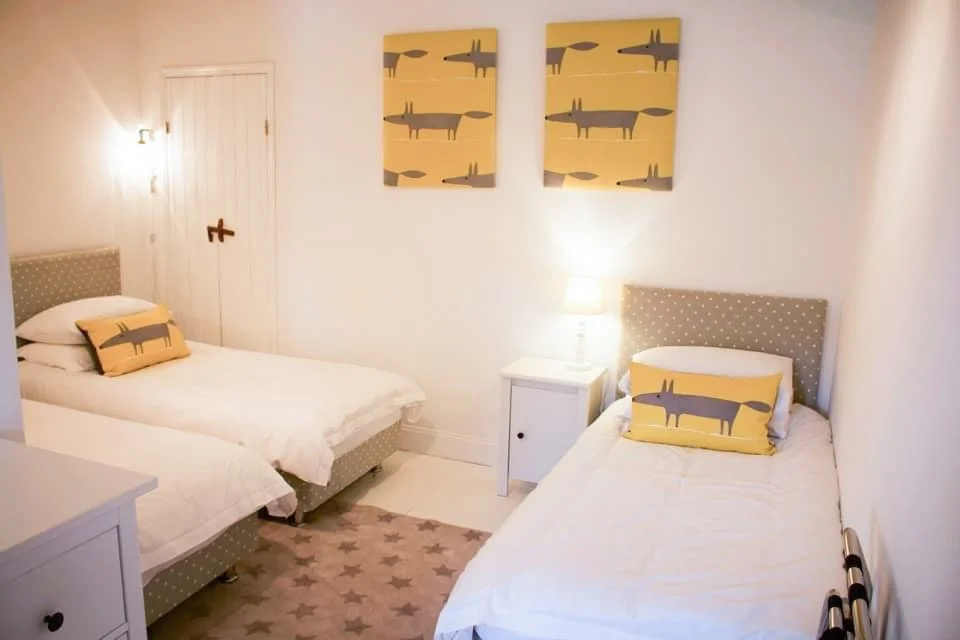 A bedroom with two white beds, each with a rectangular pillow featuring a stylized fox design in yellow and gray. There are two matching fox-themed artworks hanging above the beds, a white nightstand with a lamp between the beds, and star-patterned rugs on the floor. The room has white walls and a closed door.