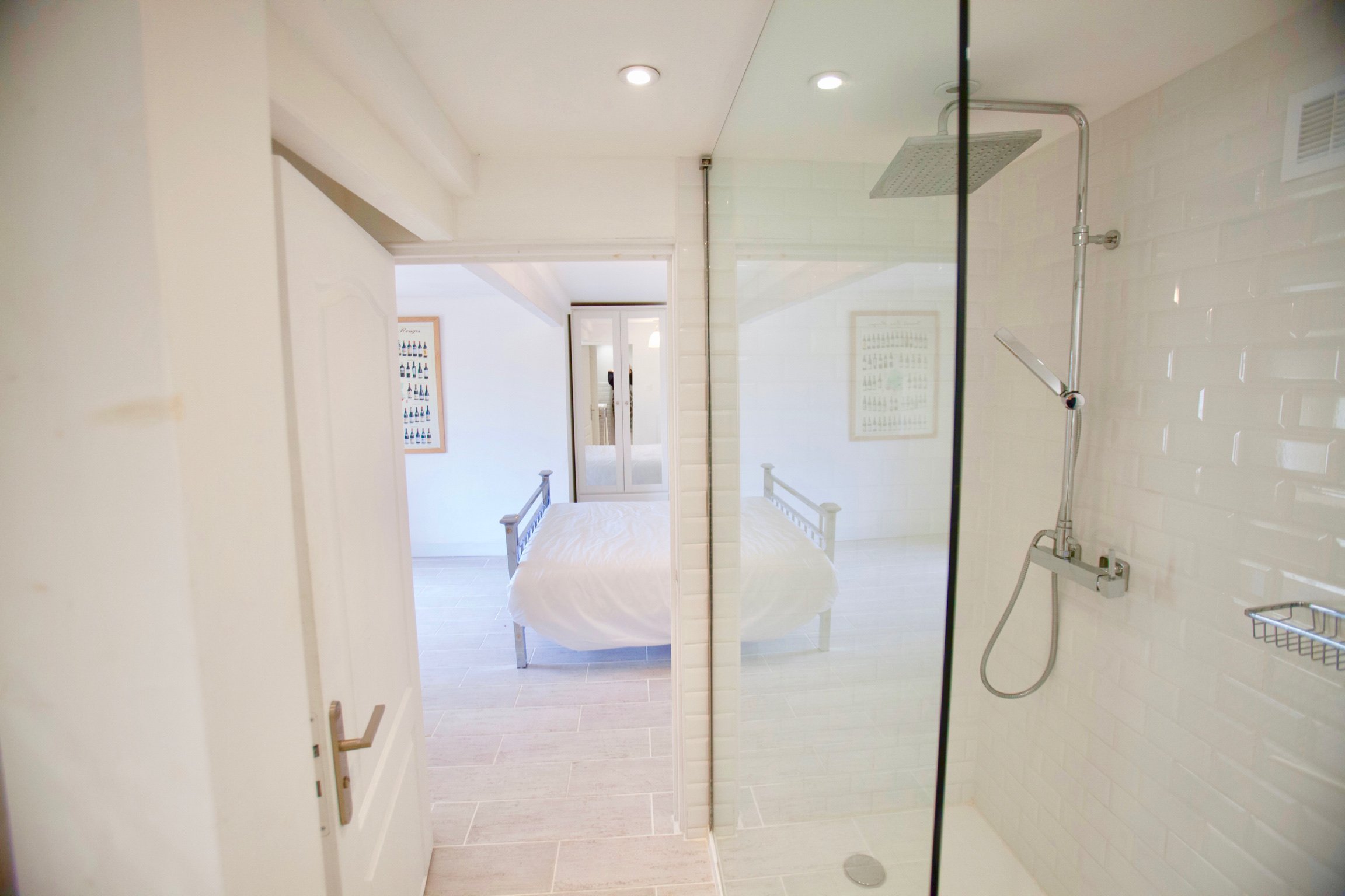 Bathroom with a glass shower, showerhead, white brick wall tiles, and a bedroom with a bed and framed artwork visible through the glass.
