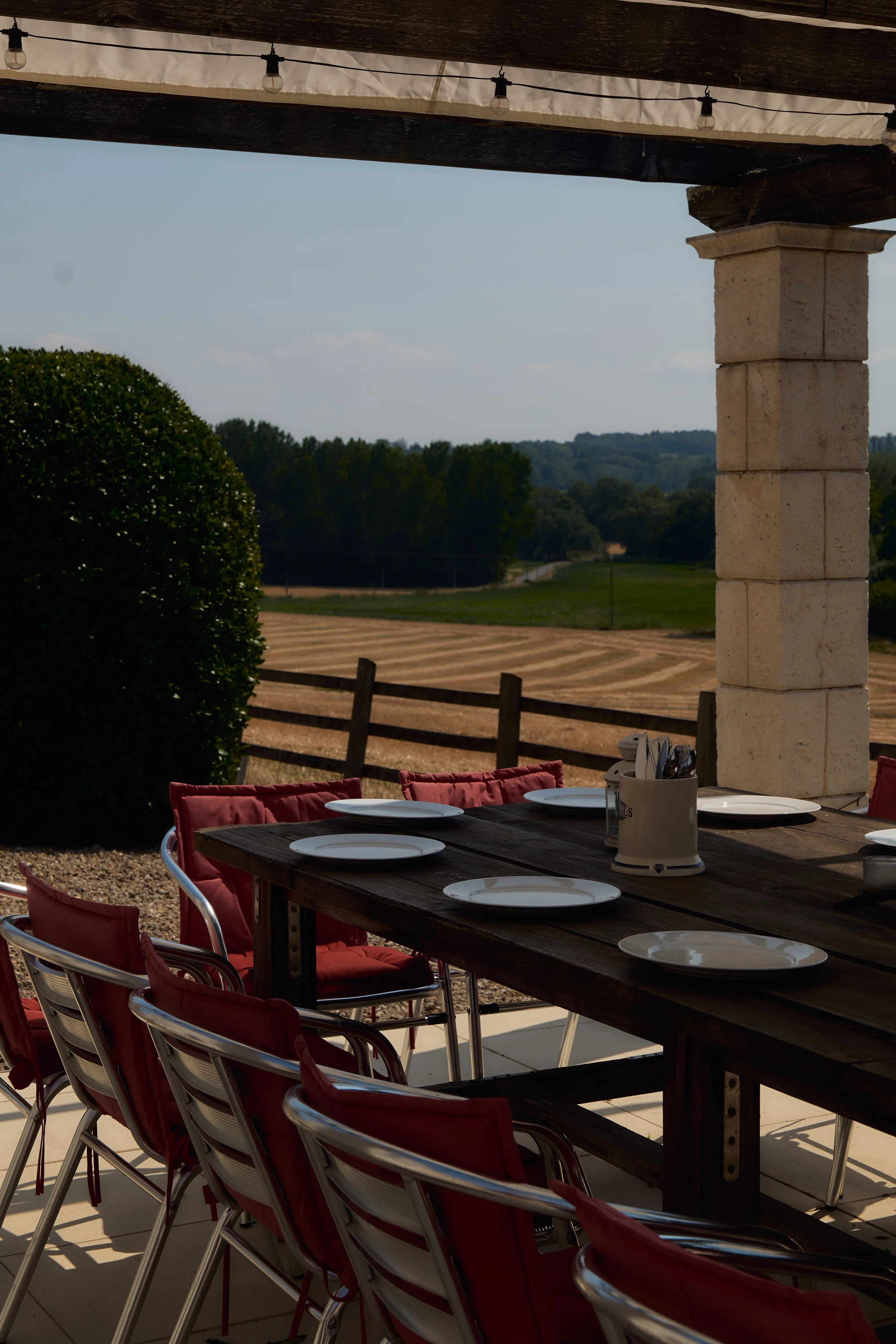 Outdoor dining table with white plates and a utensil holder, under a shaded patio with string lights, overlooking rolling fields and trees in a rural setting.
