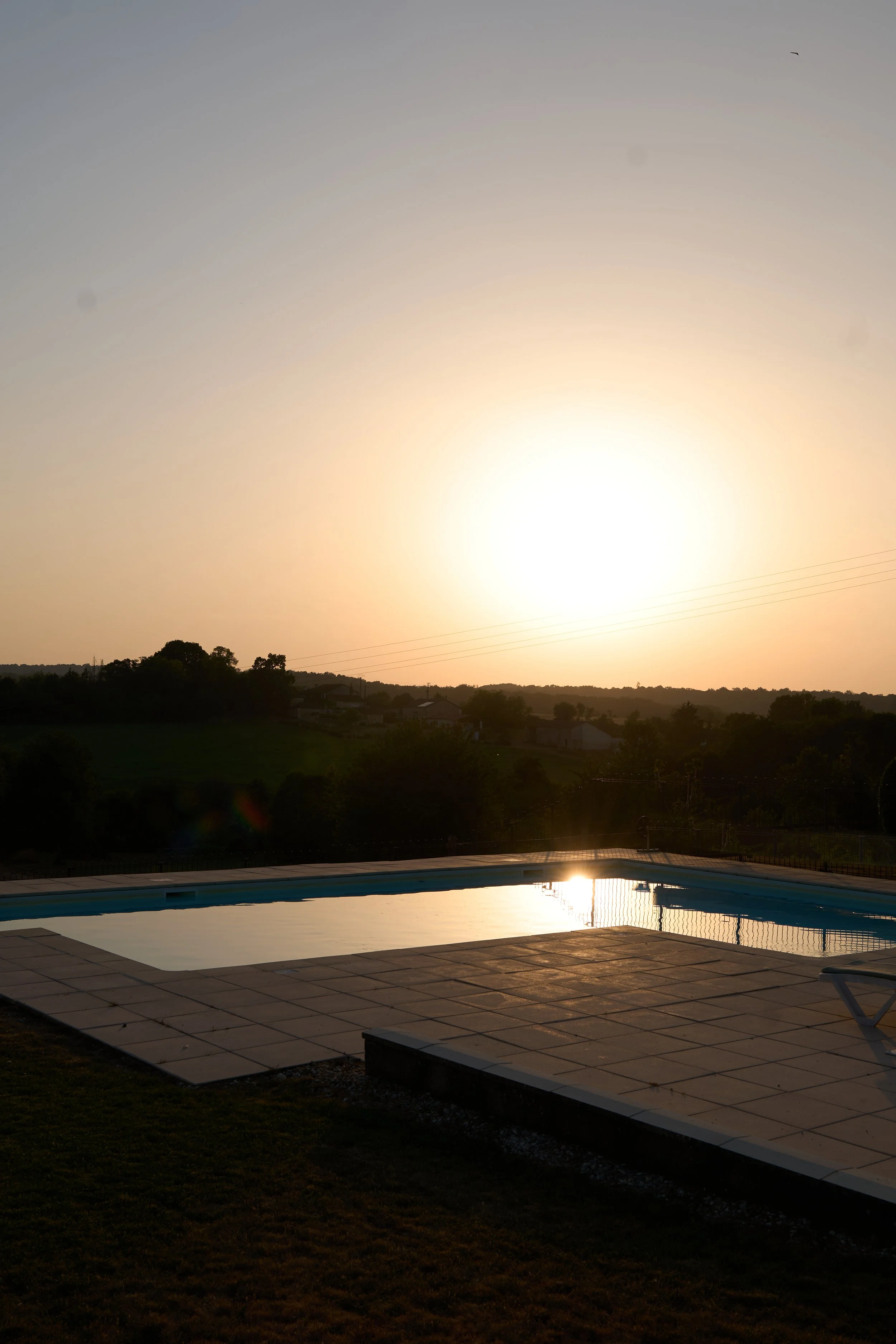 Poolside scene during sunset with a rectangular swimming pool, surrounding patio tiles, and silhouetted trees and hills in the background.