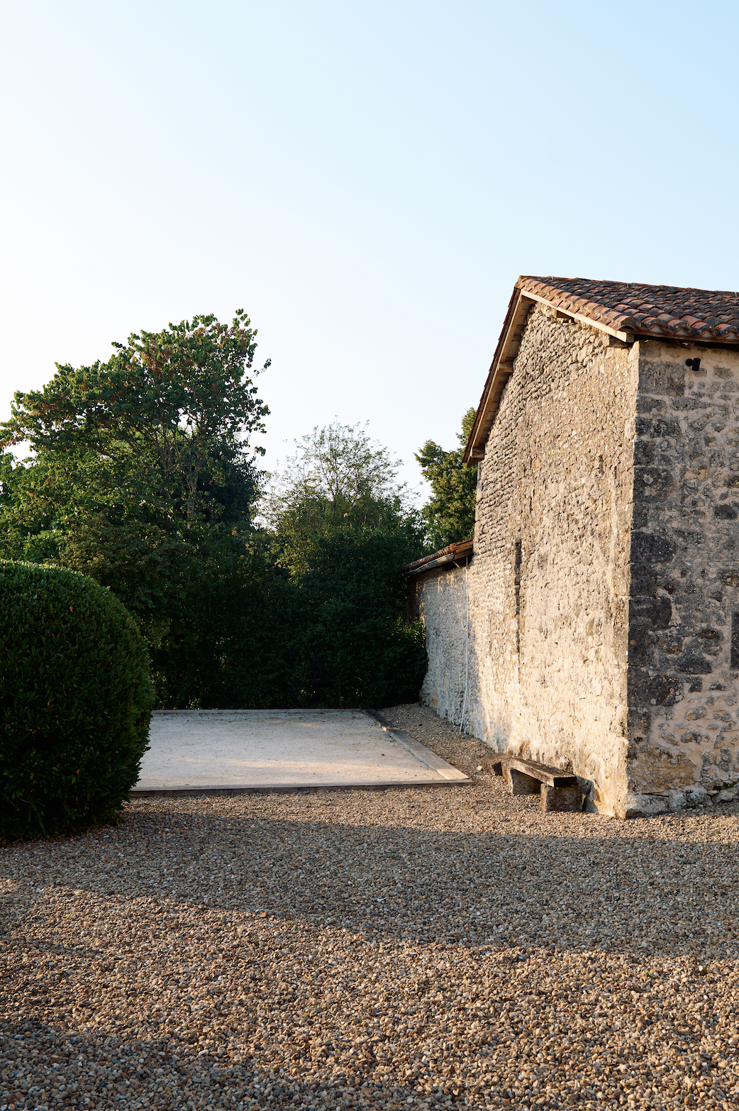 Stone building with tiled roof, surrounded by trees and gravel ground, with a small concrete area and bench.
