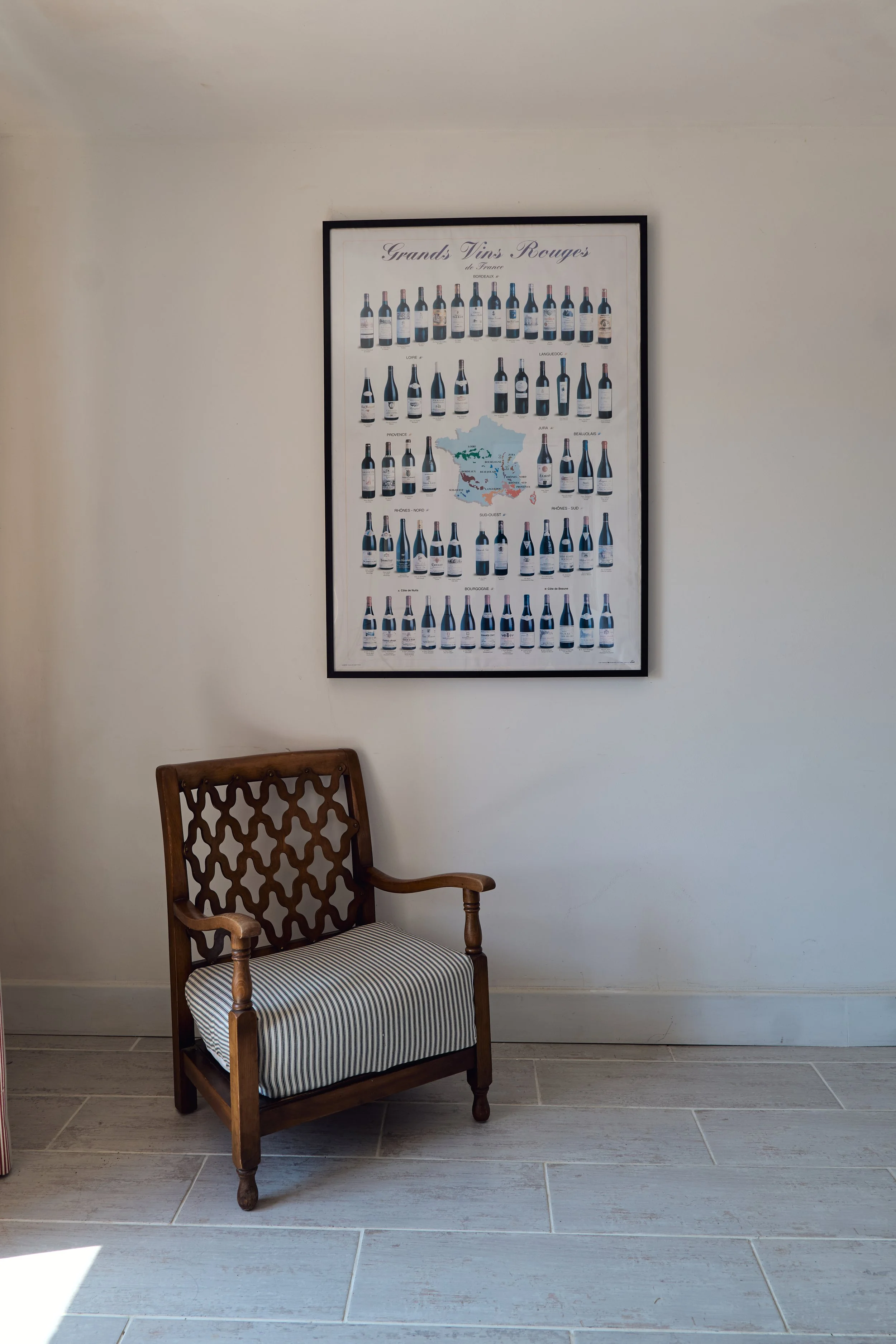 Interior scene with a wooden chair featuring patterned upholstery and a framed poster of French wine bottles on a white wall.