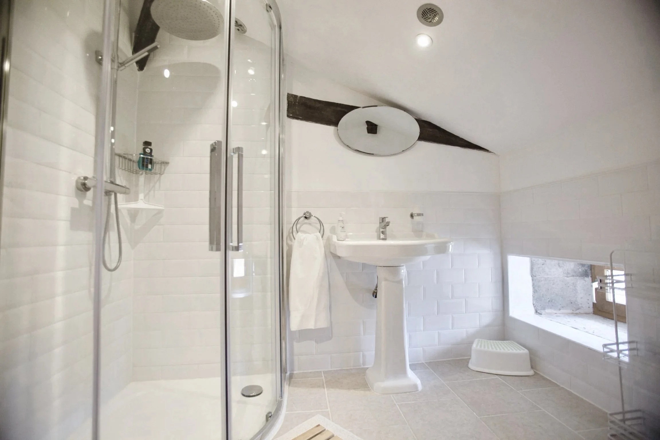 A modern bathroom with a glass-enclosed shower on the left, a white sink and mirror in the center, and a toilet to the right, all with white tiled walls and gray tile flooring, along with a small window near the ceiling.