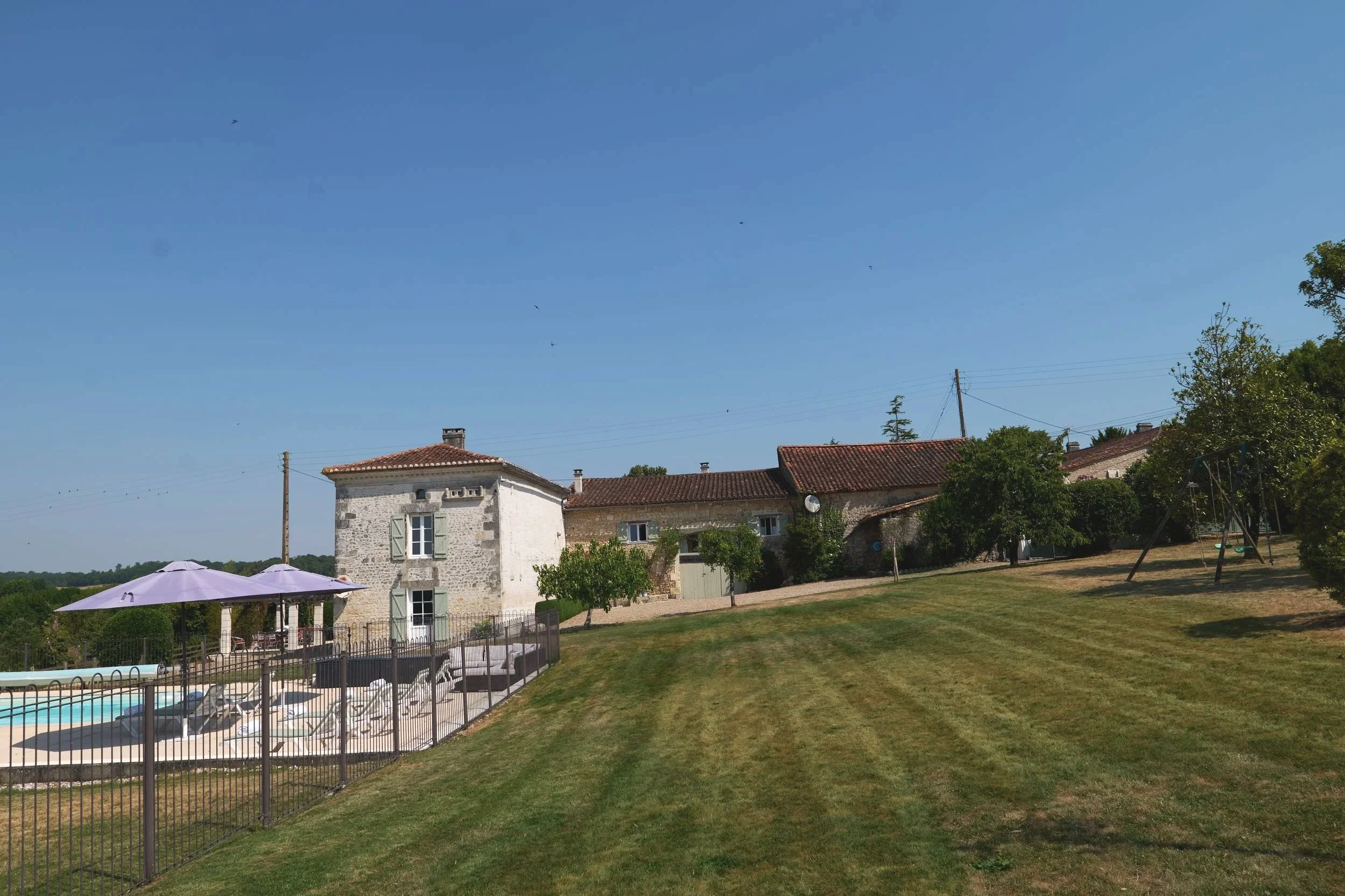 A backyard scene on a sunny day with a lawn, a swimming pool area with umbrellas, a stone house with green shutters, and trees under a clear blue sky.