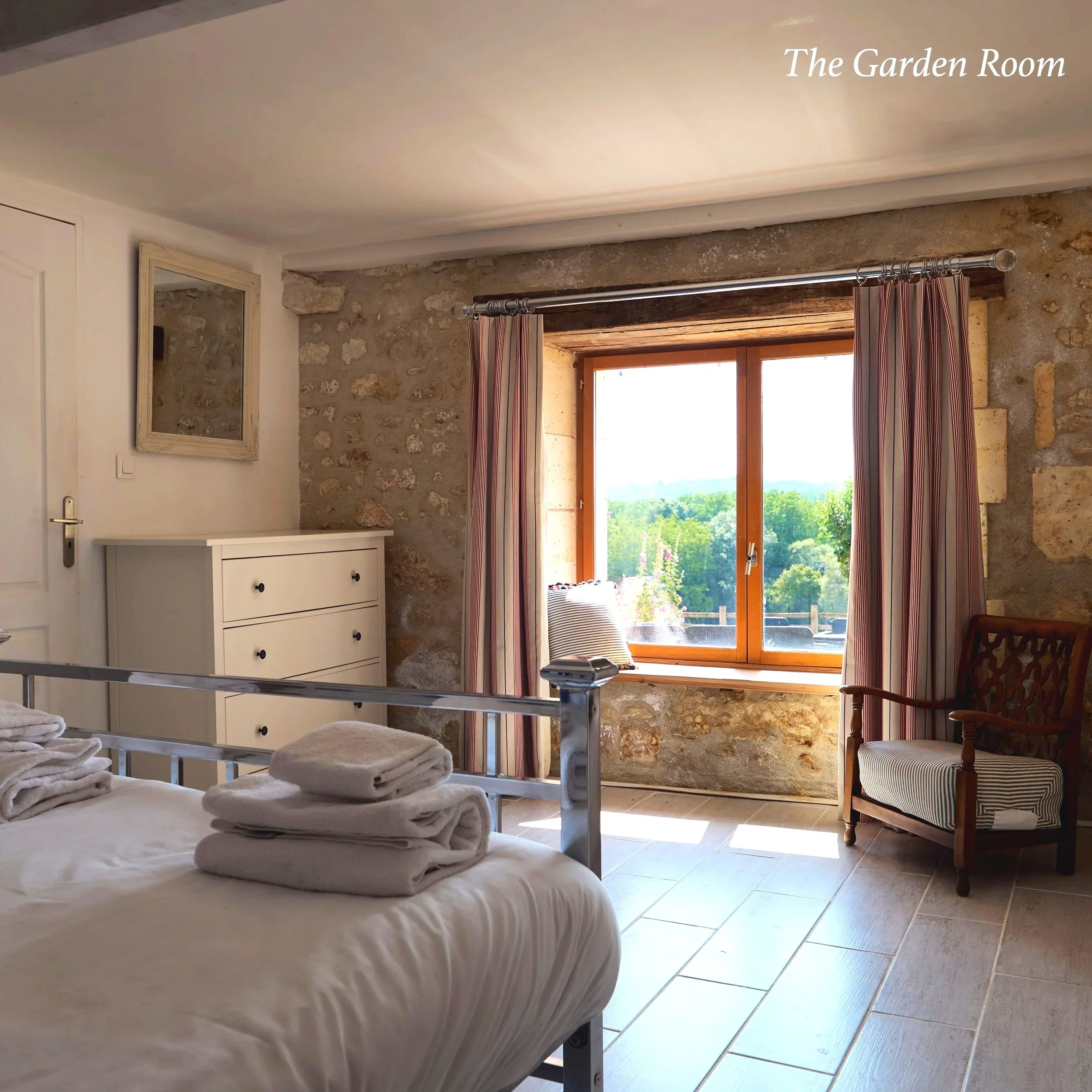 The image shows a cozy bedroom with a stone wall, a large window with striped curtains, a wooden chair, and a white dresser with a mirror. The bed has white linens and towels folded on top.