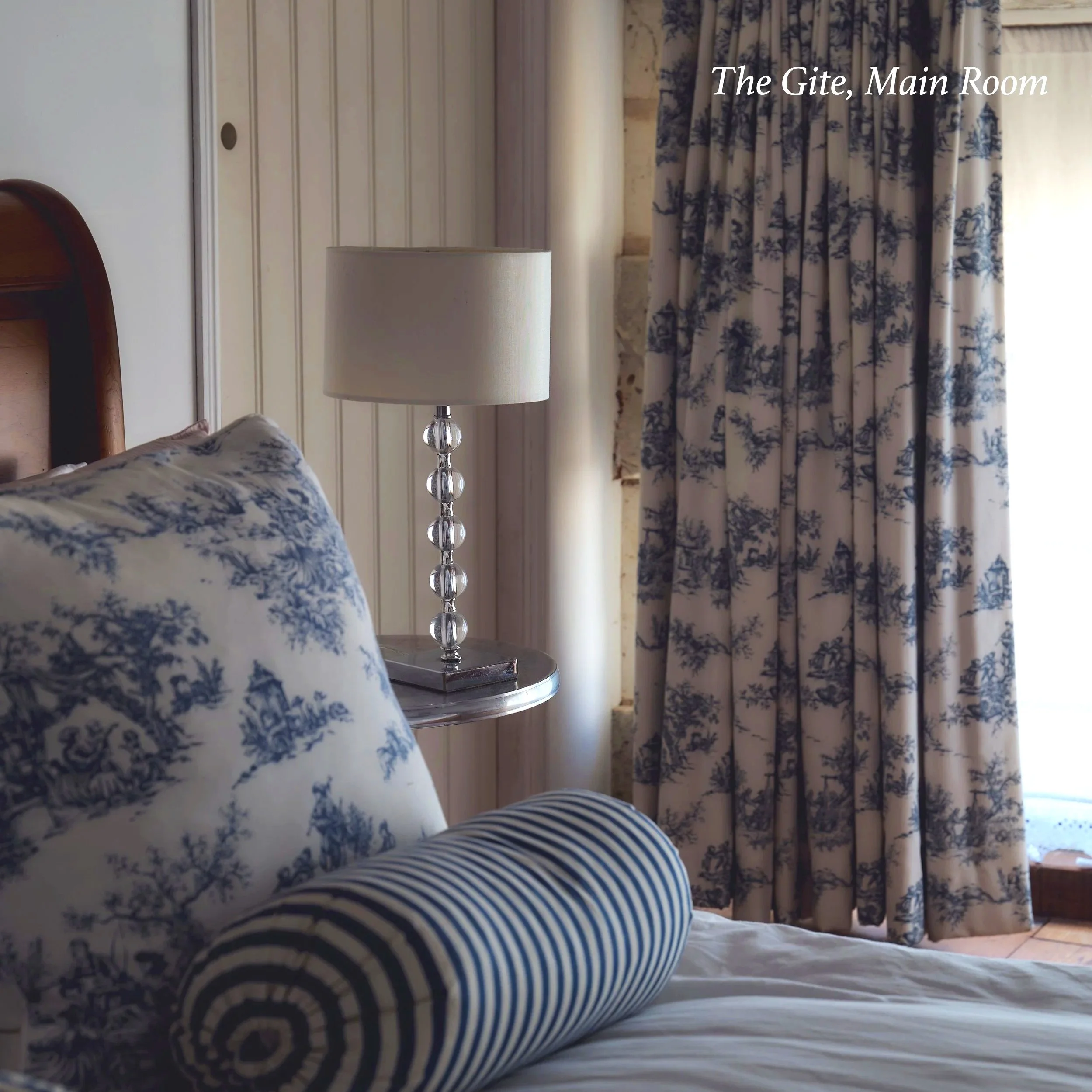 A bed with patterned pillows and a bedside lamp, partly covered by blue and white striped bedding in a cozy room with floral curtains and wooden wall paneling, in a hotel or guesthouse setting.