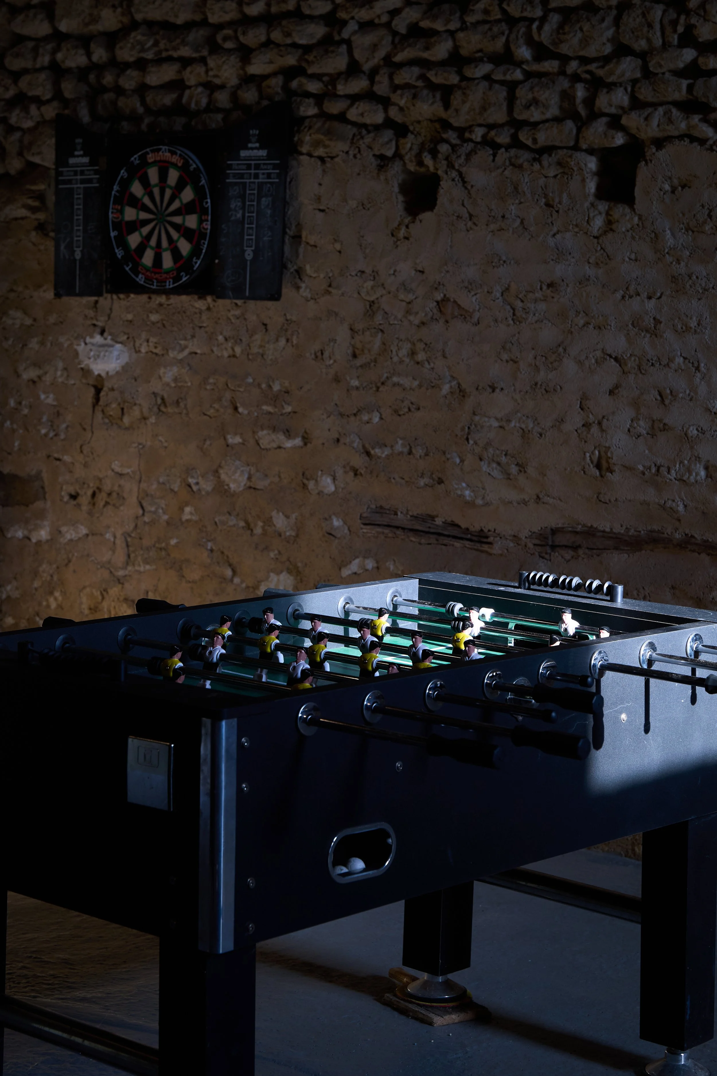 A foosball table in a dimly lit room with a dartboard on a textured, brown wall in the background.