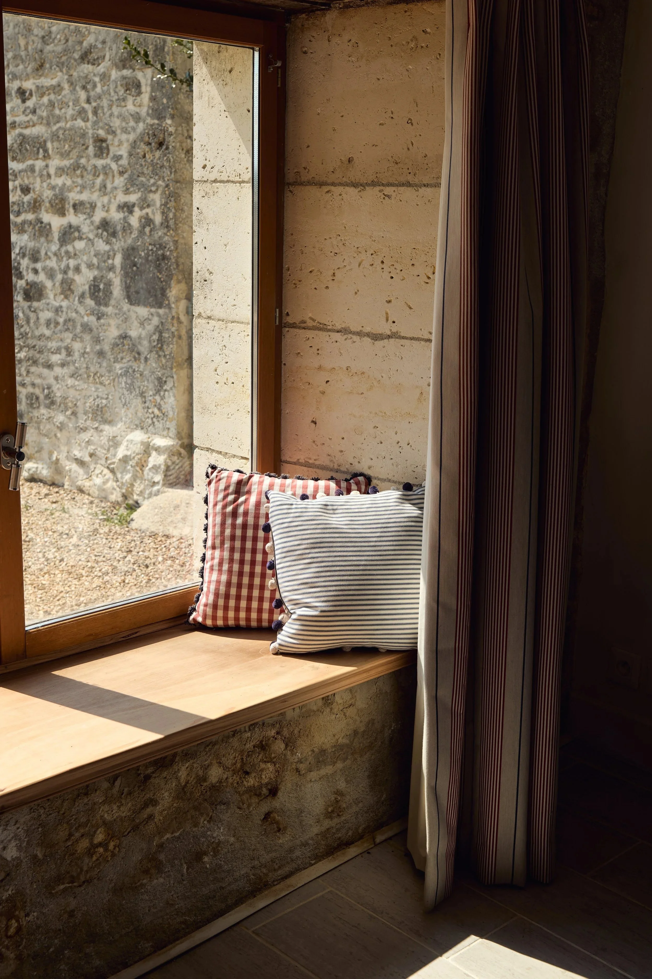 A windowsill with two striped pillows, window with a stone wall outside, and a striped curtain partially drawn. Sunlight is casting shadows on the floor.