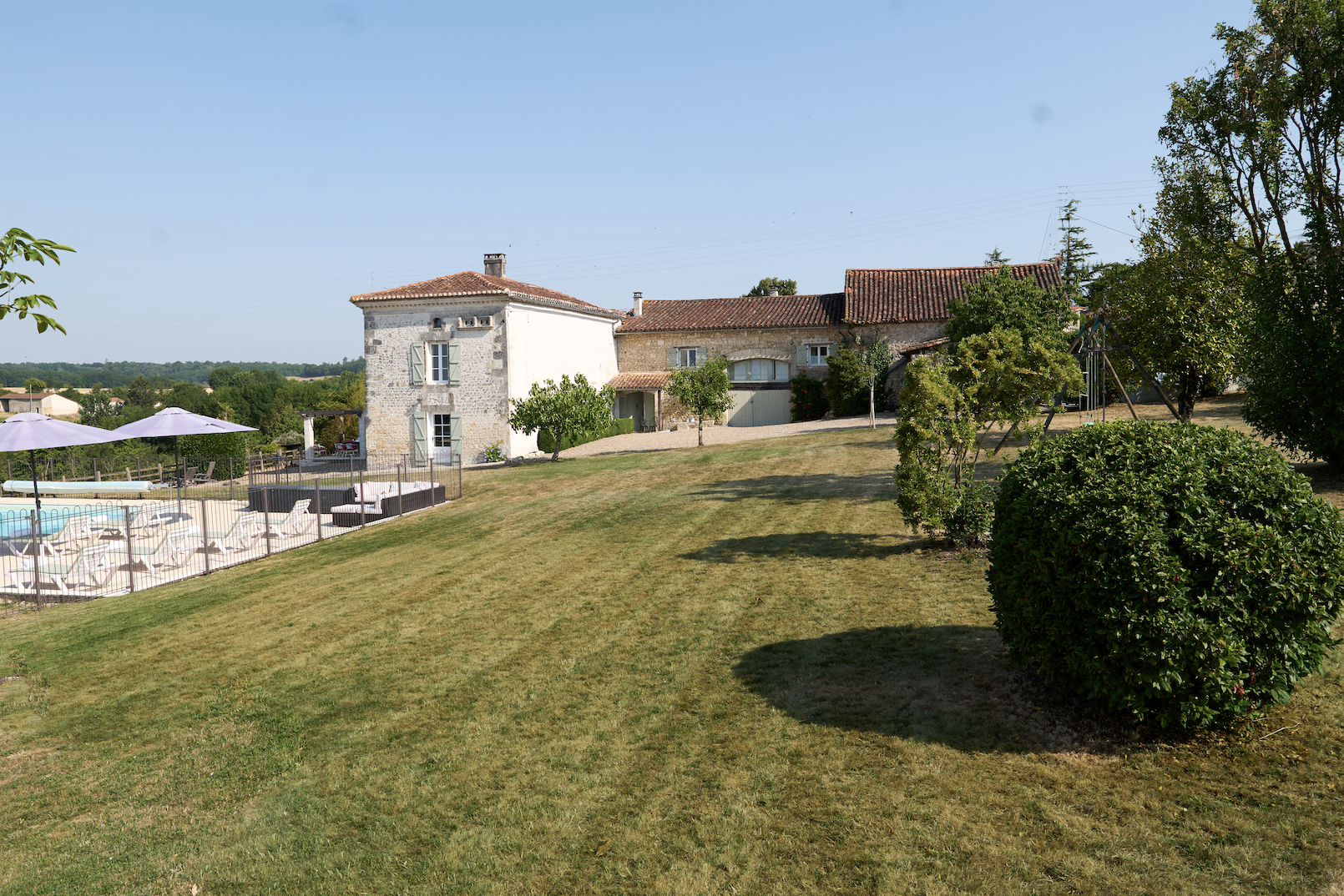 A rural house on a hill with green lawn, trees, and a swimming pool with umbrellas and lounge chairs, under a clear blue sky.