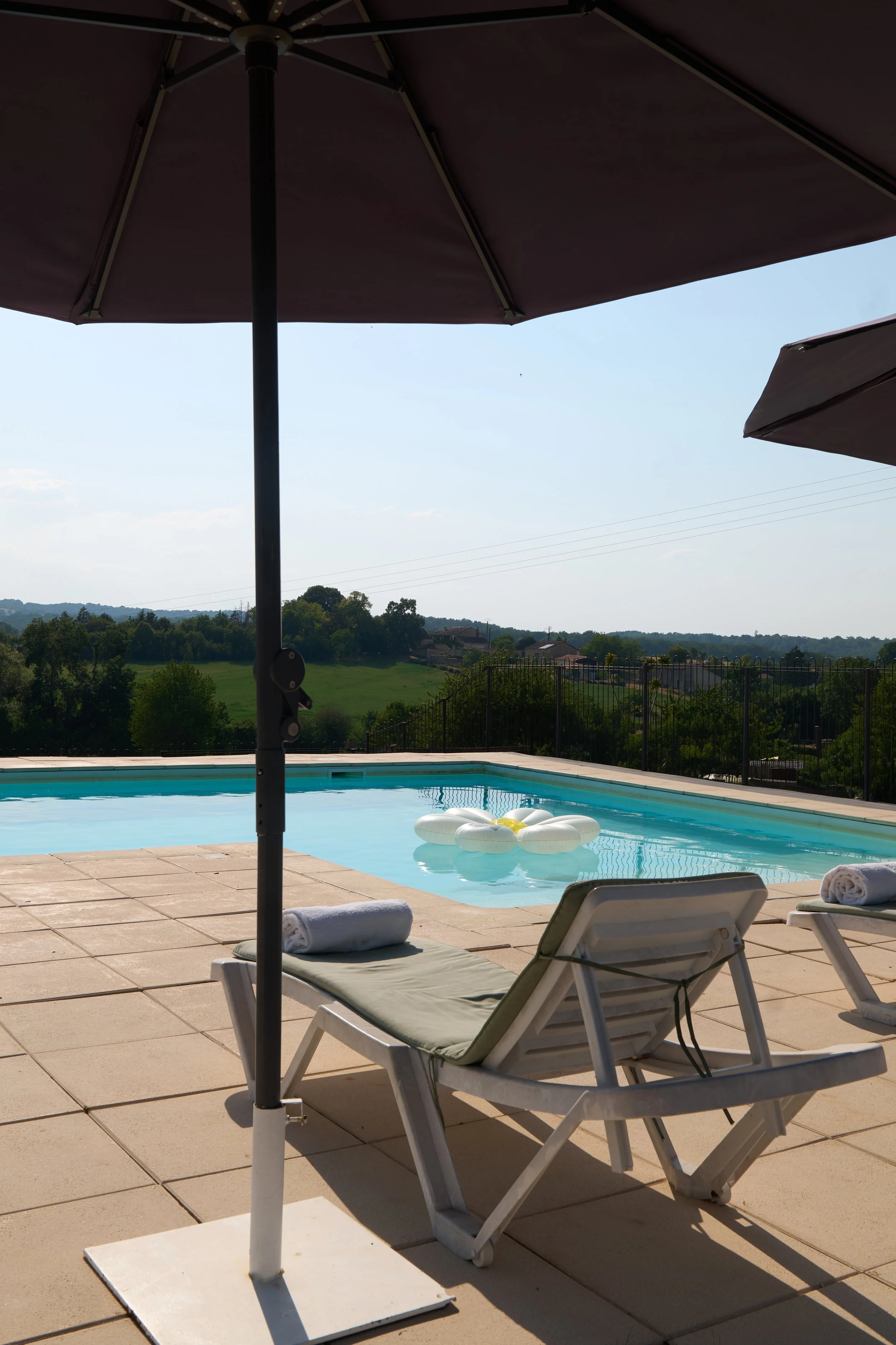 Poolside scene with lounge chairs, rolled towels, a large umbrella, and a swimming pool, overlooking a green landscape and blue sky.