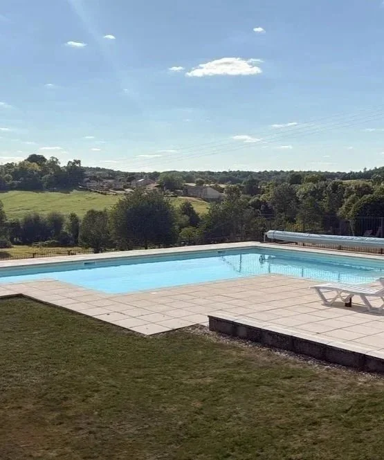 Outdoor swimming pool with a lounge chair on a tiled deck, green grassy yard, rolling hills in the background, and a blue sky.
