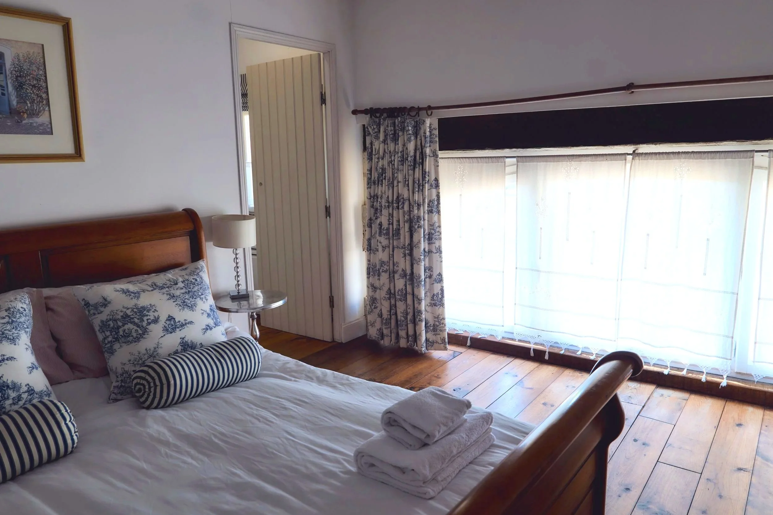 A bedroom with a wooden bed, white bedding, and decorative pillows. Two rolled towels are placed on the bed. Next to the bed, there is a bedside table with a lamp. The room has large windows with sheer white curtains and patterned drapes, allowing sunlight to brighten the space. A picture hangs on the wall near the door, which is slightly ajar, revealing another room.