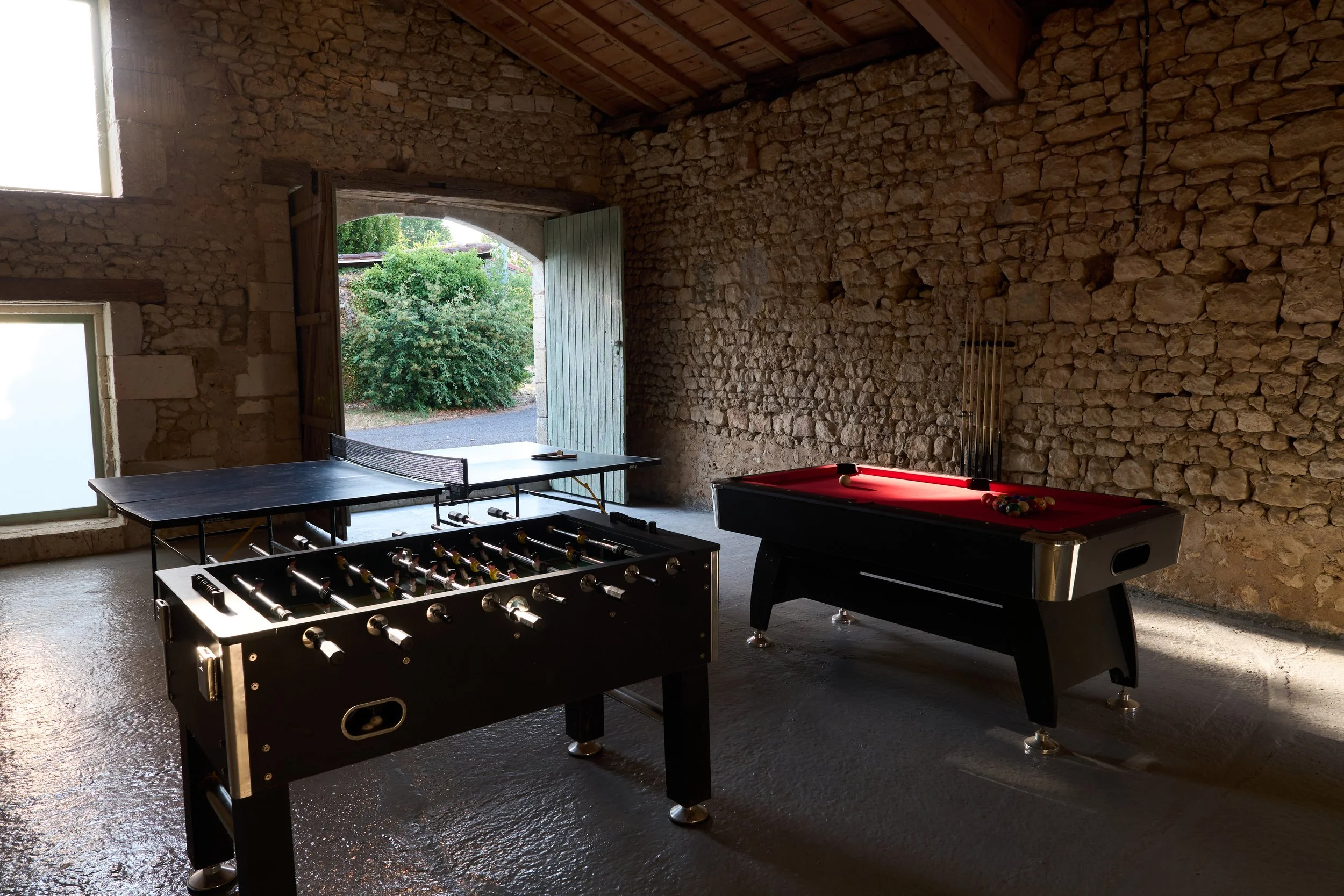 An indoor game room with stone walls and wooden beams, featuring a foosball table, a pool table with a set of balls on it, a table tennis table, an open door showing greenery outside, and a window letting in light.