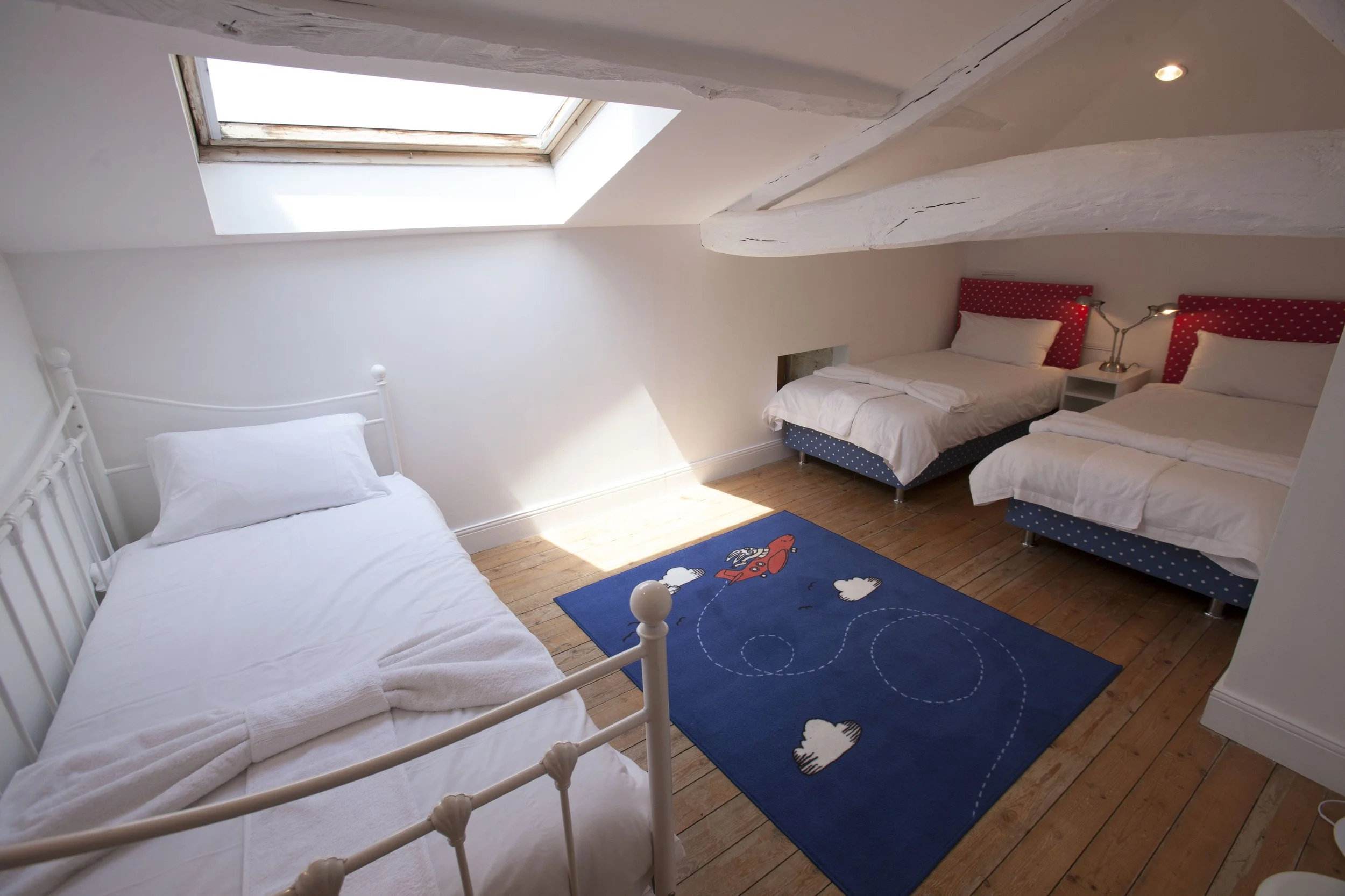A bedroom with two beds, one with a white iron frame and the other with a colorful headboard with red and blue polka dots, a skylight window, a blue rug with cloud and rocket designs, and wooden floors.