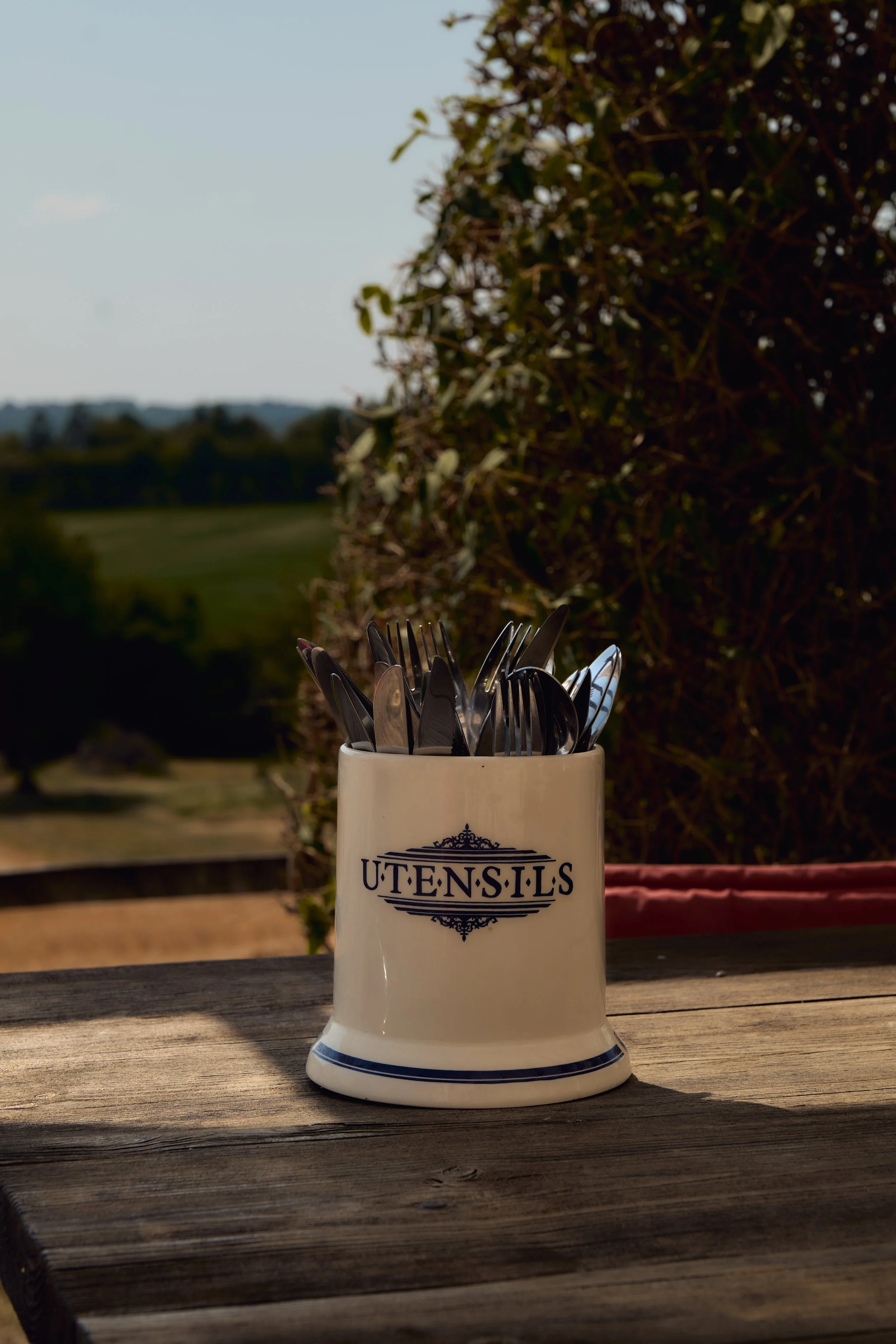 A white utensil holder with the word 'UTENSILS' printed on it, containing silver forks and knives, situated on a wooden table with an outdoor background featuring greenery and hills.