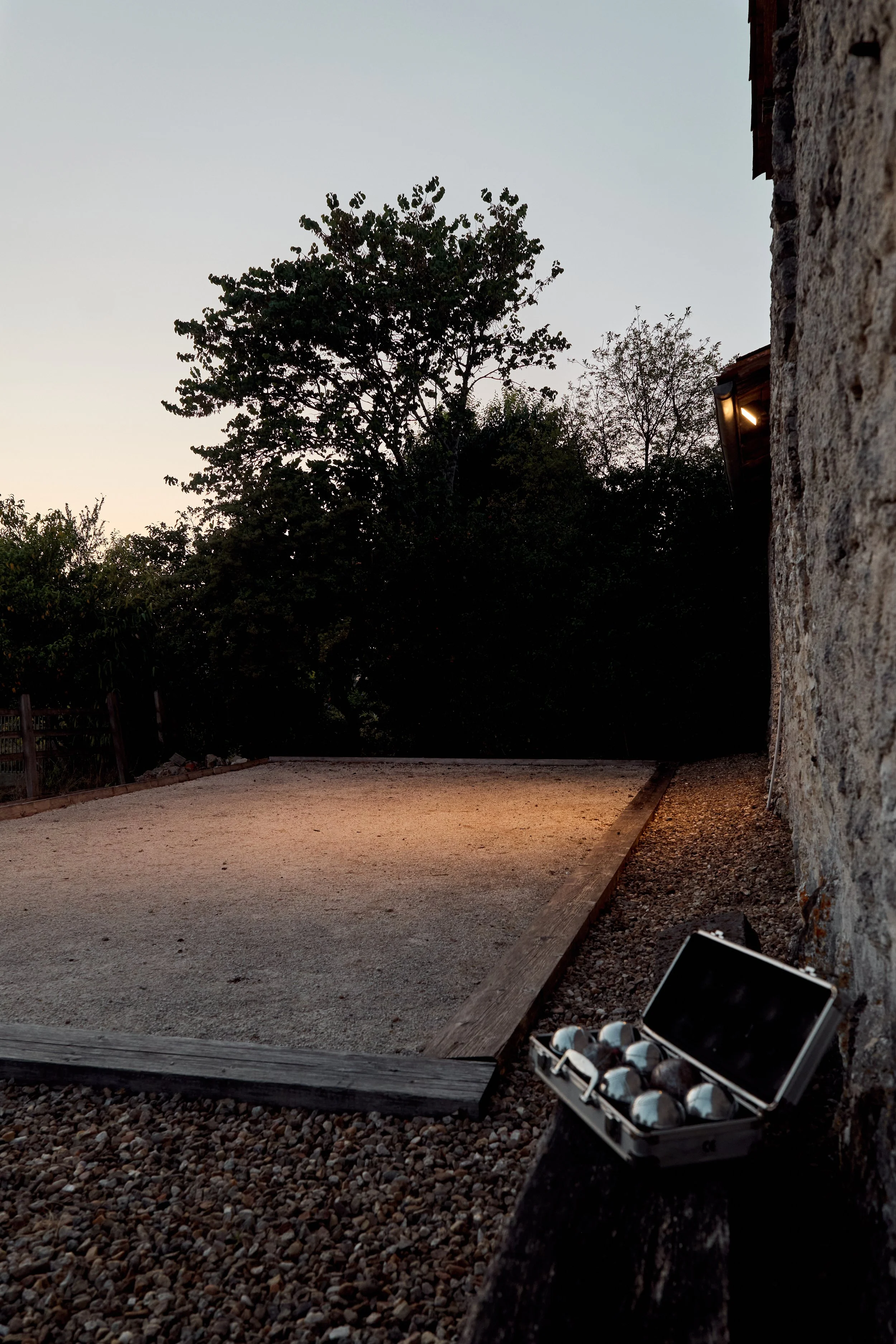 An outdoor scene at dusk showing a gravel pathway next to a stone wall, with trees in the background and an open toolbox filled with golf balls in the foreground.