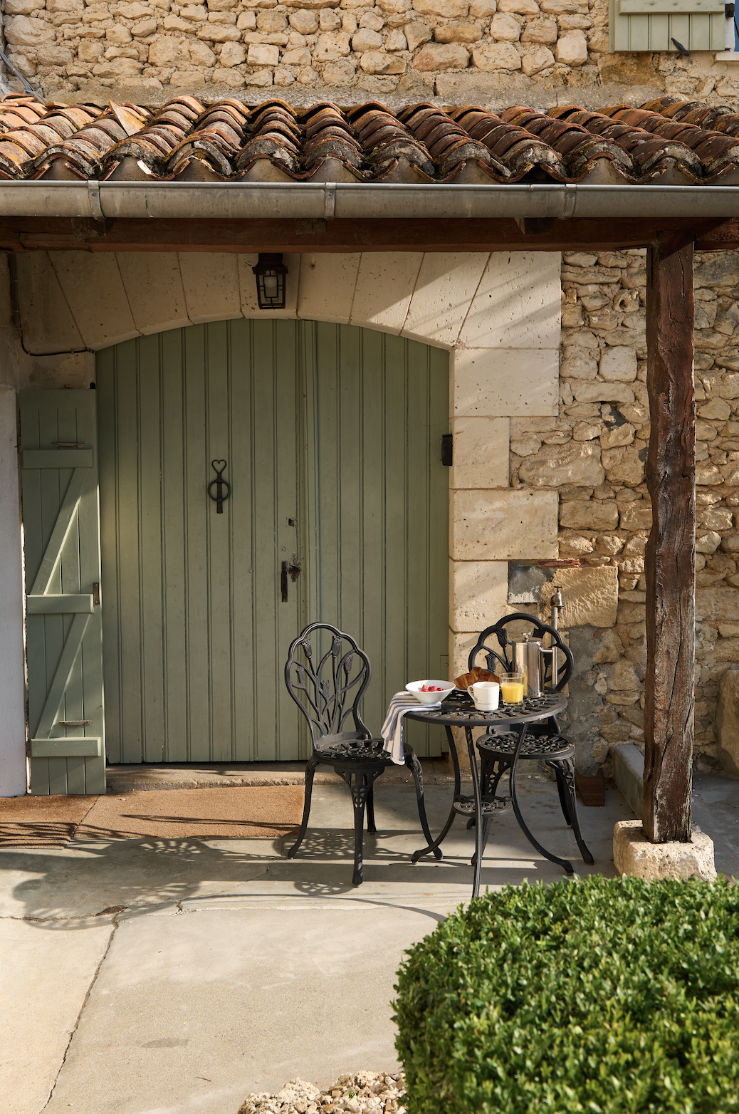 A small outdoor breakfast scene with a black metal table and two matching chairs in front of a stone building with a green arched door. The table has a breakfast set including a bowl of berries, a croissant, a cup, a glass of orange juice, and a coff