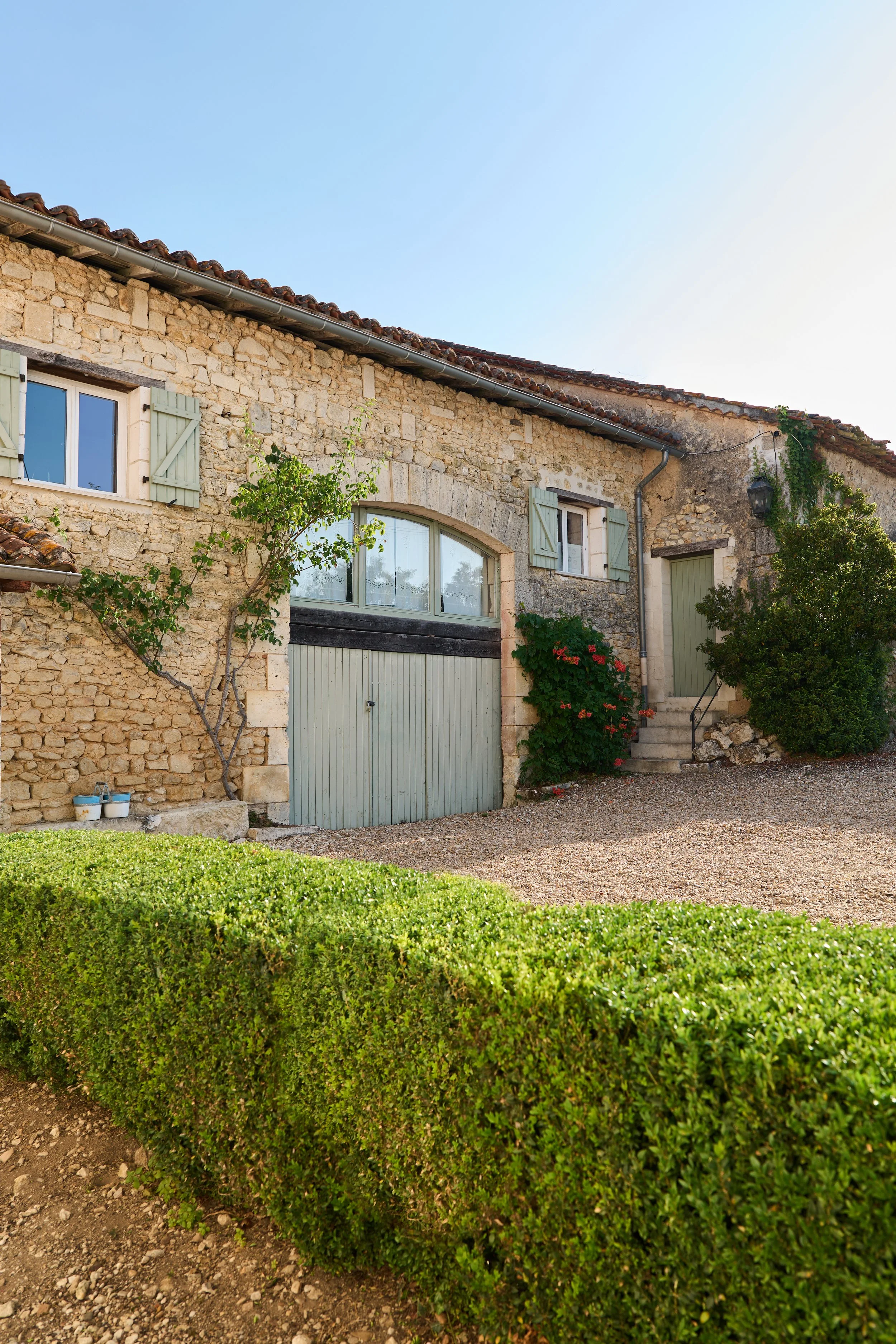 Stone house with green shutters, large garage door, stone stairs, and shrubs in front, under a clear sky.