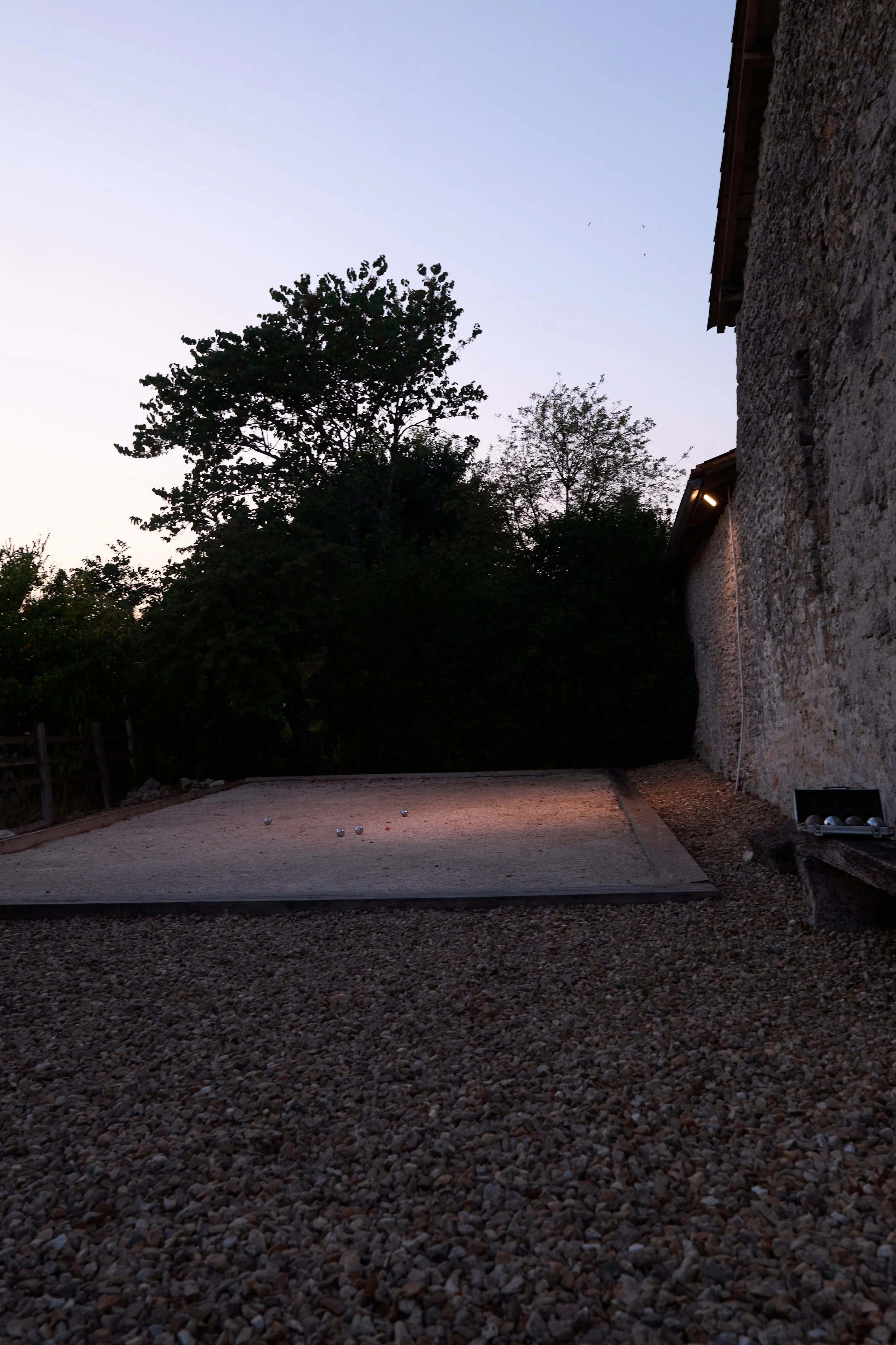 An outdoor petanque court with several metallic balls on the gravel surface, adjacent to a stone building, during twilight with trees in the background.