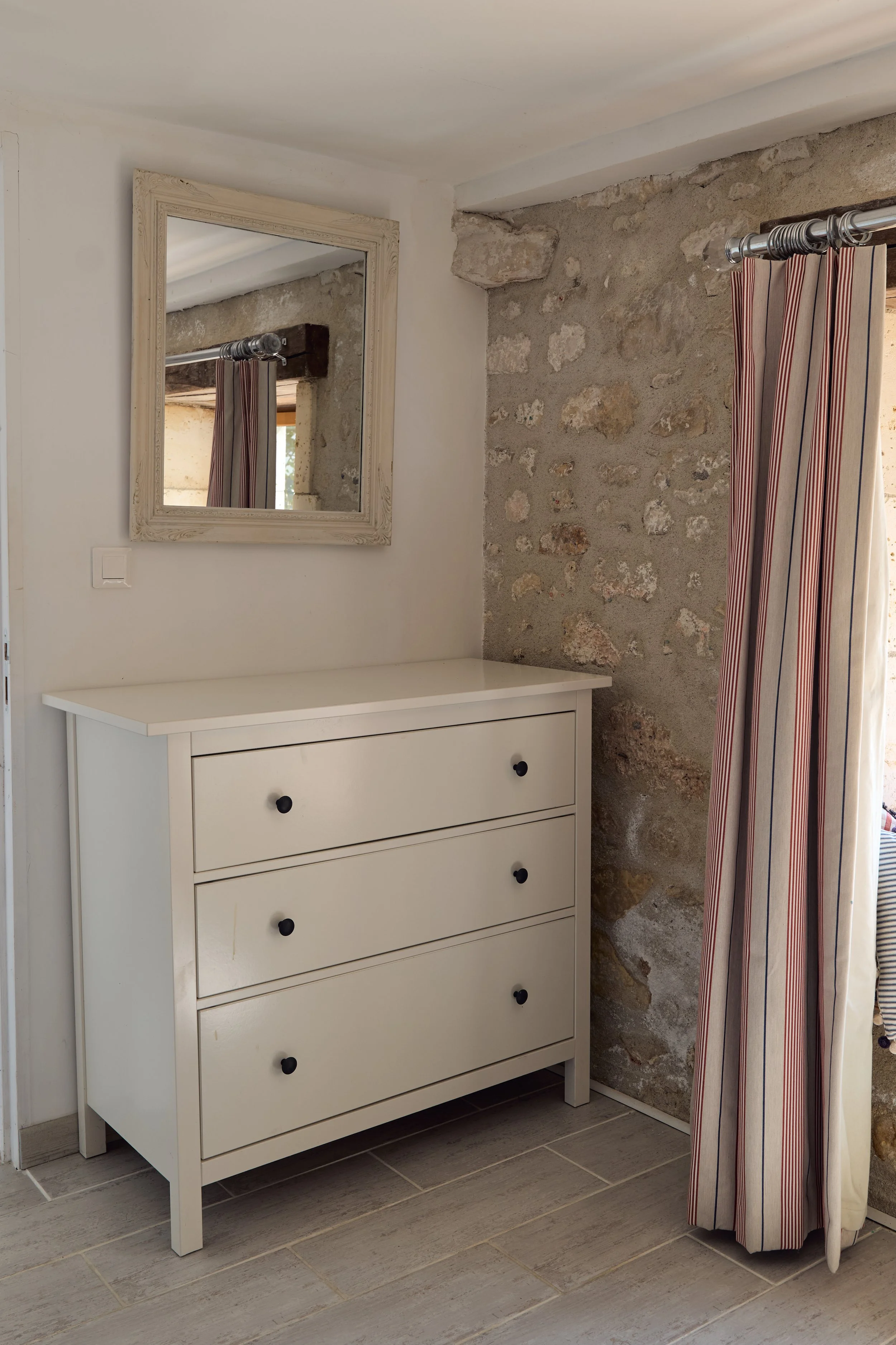 A white dresser with six drawers and black knobs in a room with a stone wall and a mirror on the wall. There is a striped curtain on a rod beside the dresser.