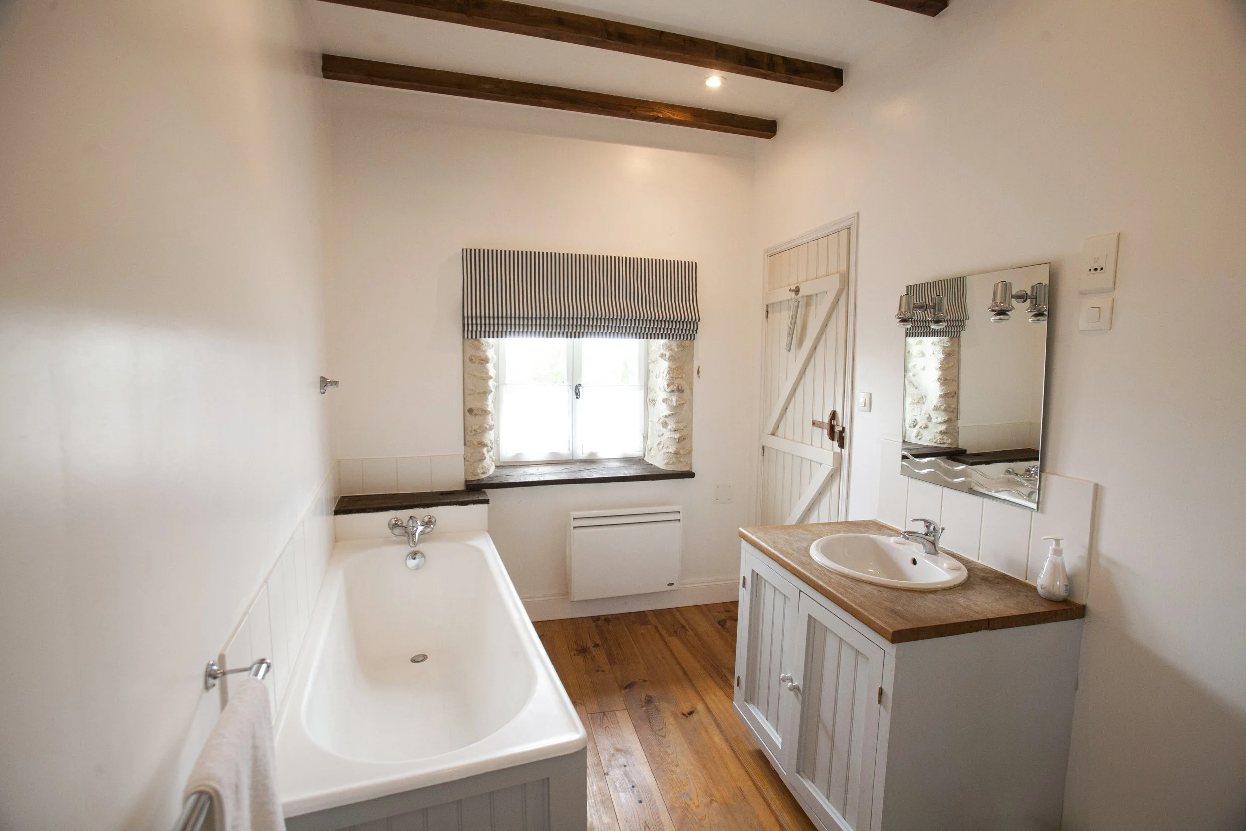 A small bathroom with a bathtub, a window with a striped valance, a mirror, a sink with a wooden countertop, and a white door with a barn-style latch. The ceiling has exposed wooden beams.