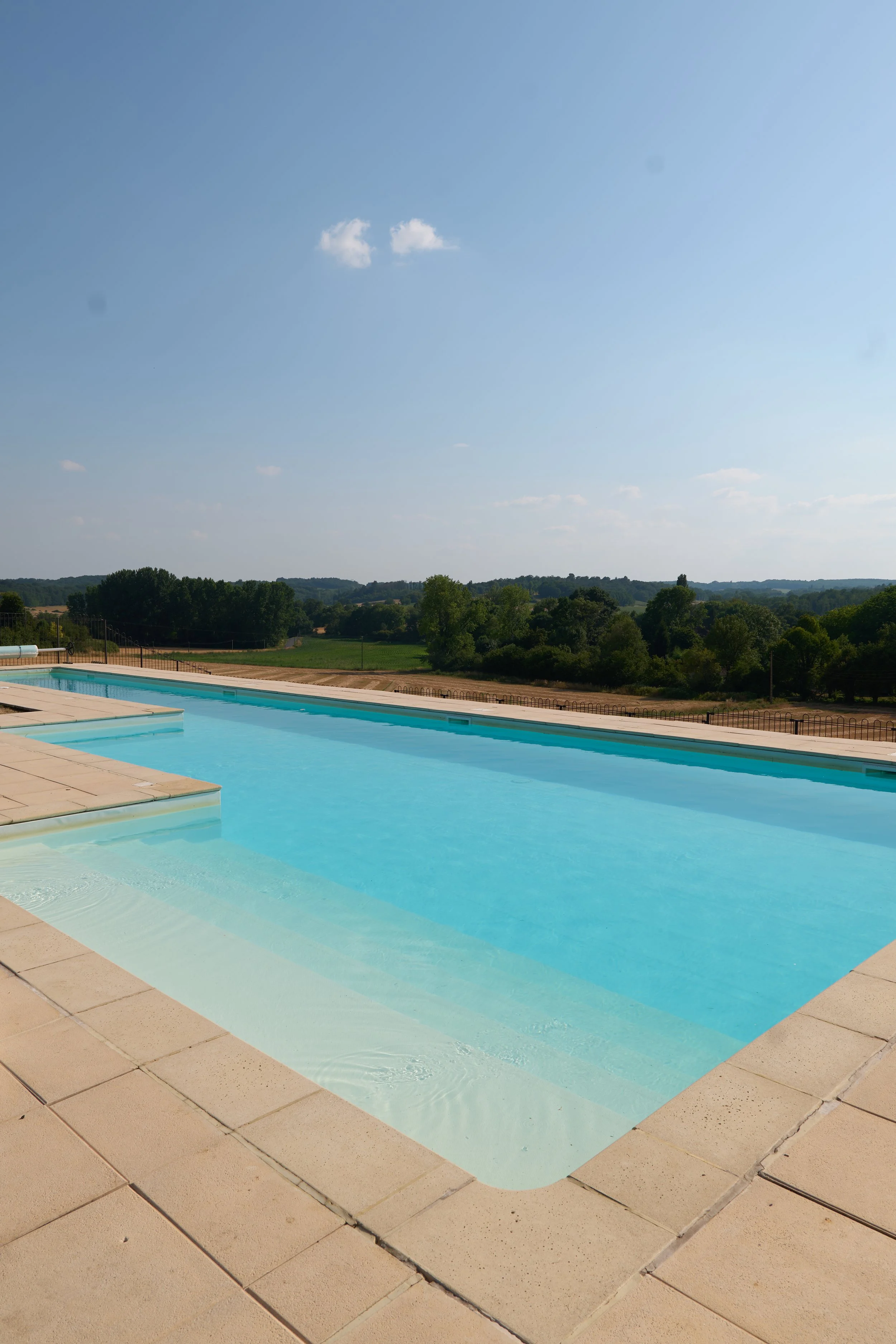 Swimming pool with beige tile deck, surrounded by a rural landscape with trees and hills under a clear blue sky.