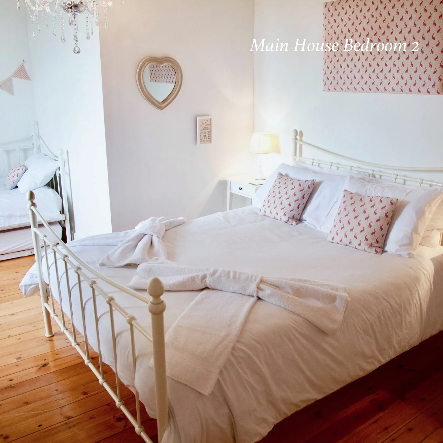 A bedroom with a white metal bed frame, white bedding, and three pillows with pink heart patterns. There is a small white nightstand with a lamp on it. The wall behind the bed has a pink heart-patterned artwork and a decorative hearts mirror. The flo