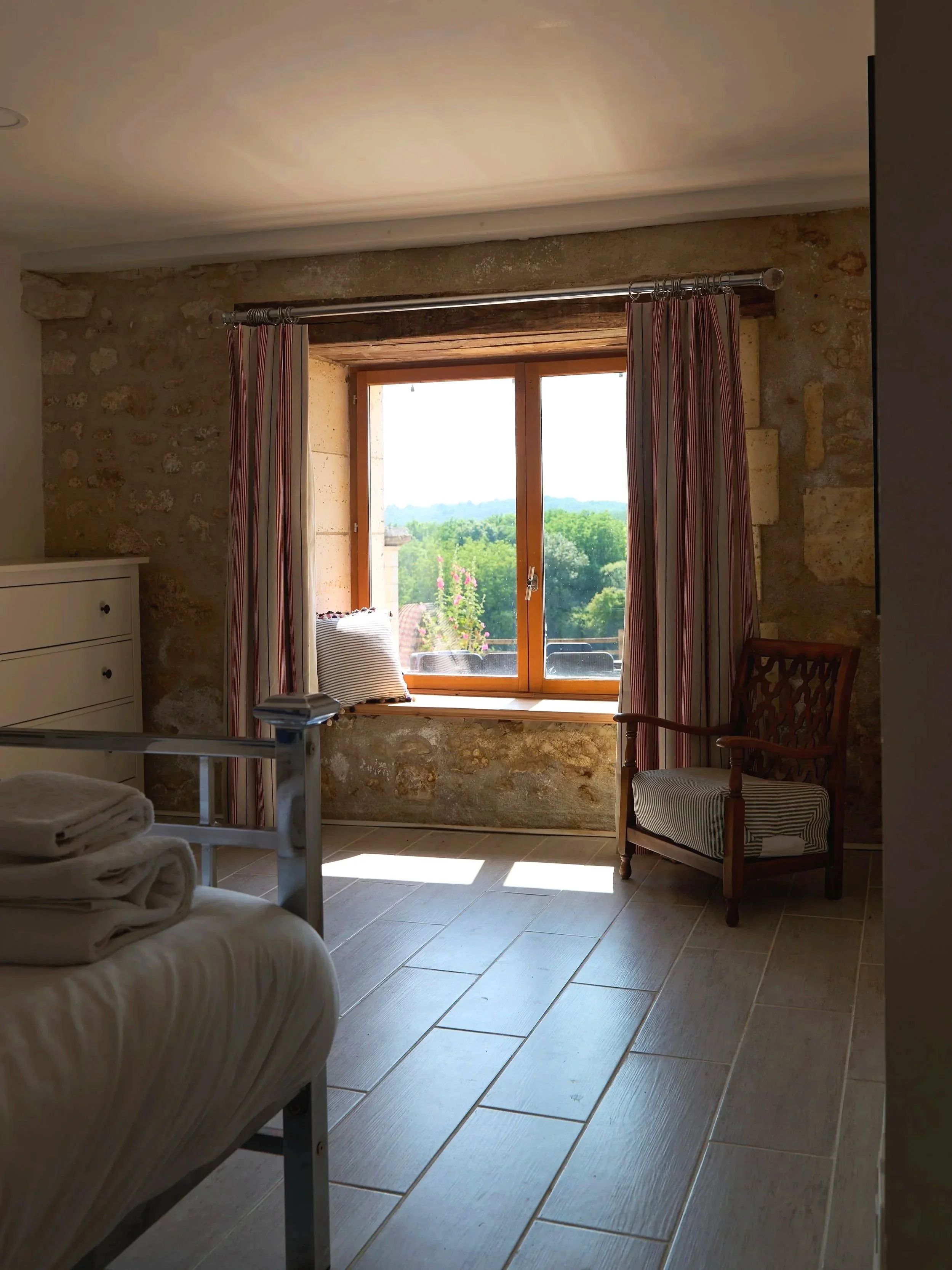 Sunlight illuminating a rustic bedroom with a window view of greenery, a chair, and a bed with folded towels.