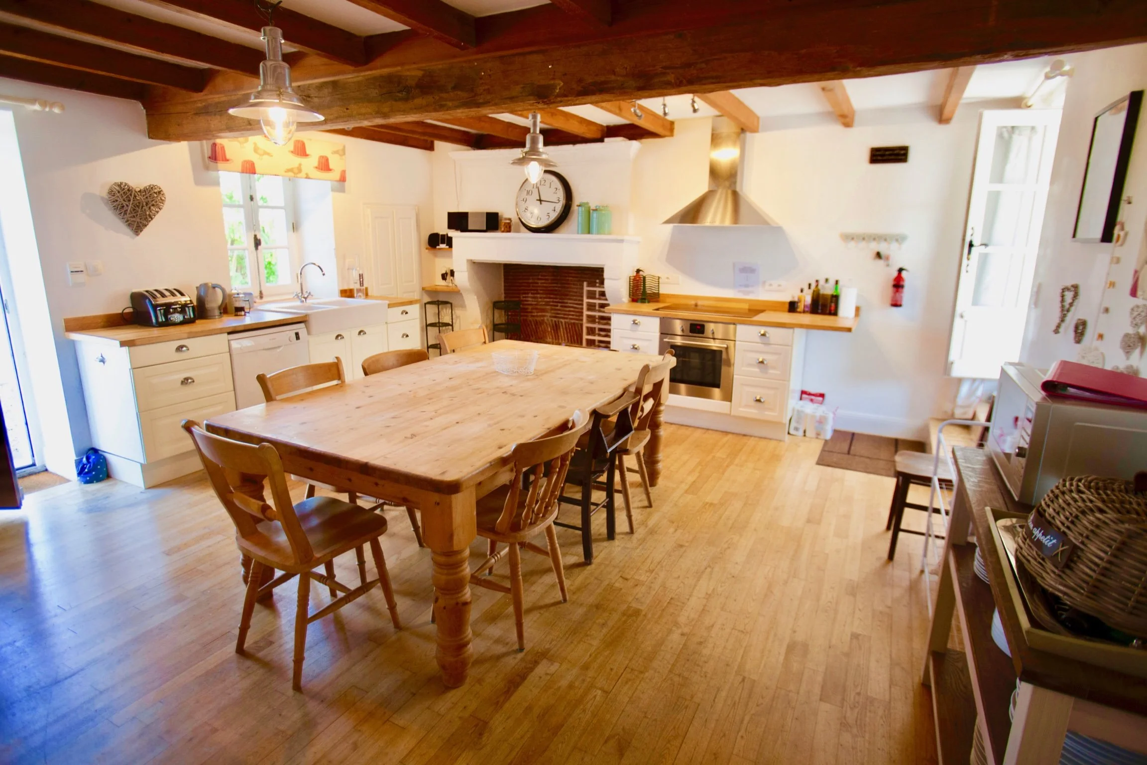 A cozy kitchen with wooden beams on the ceiling, a large wooden dining table with chairs, white cabinetry, and a fireplace with a clock above it. There is a window with a decorative valance and various kitchen appliances and decor items.