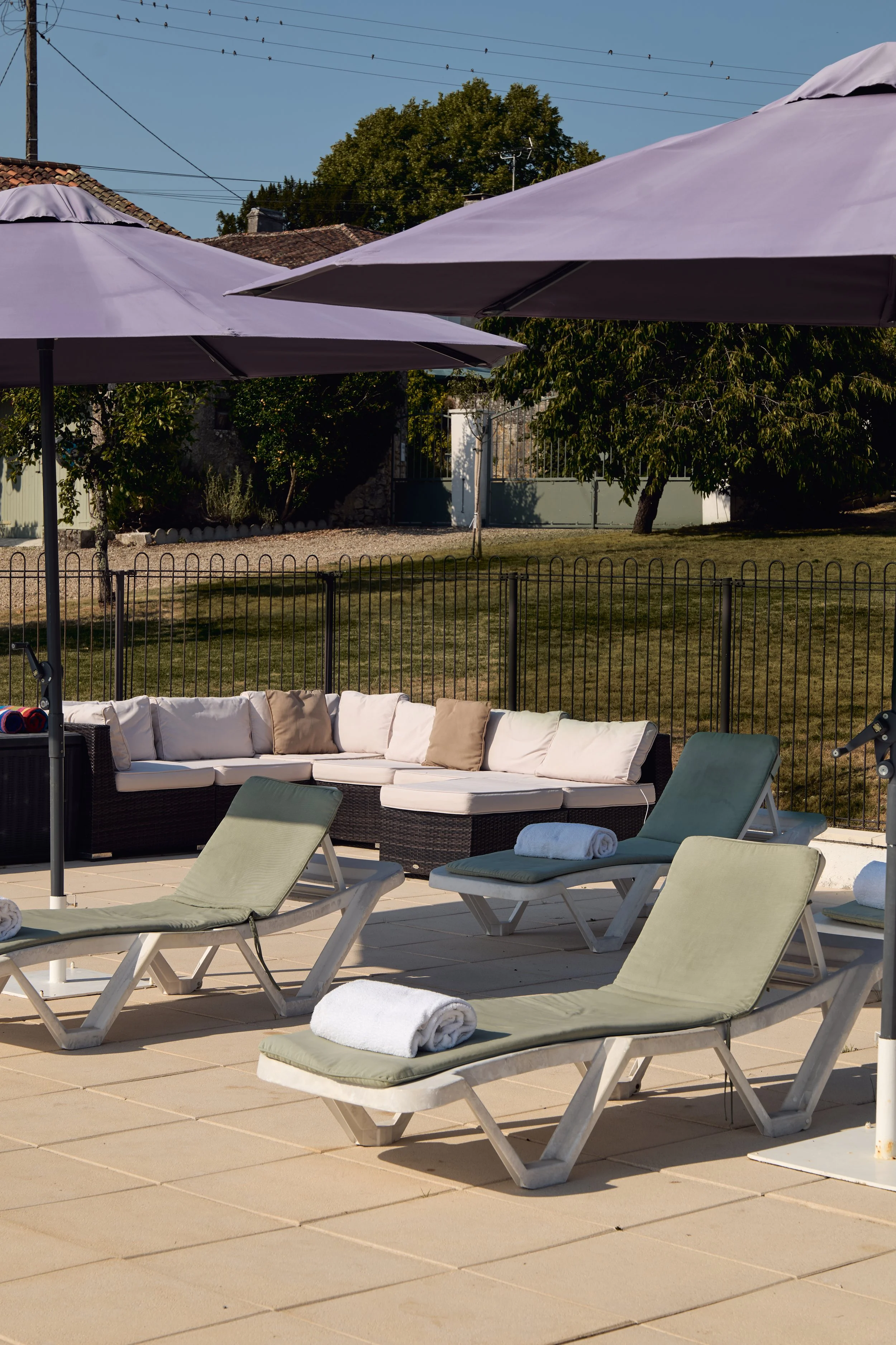 Outdoor lounge area with green sunbeds, beige cushioned seating, two large purple umbrellas, and a fenced backyard with trees and houses in the background.