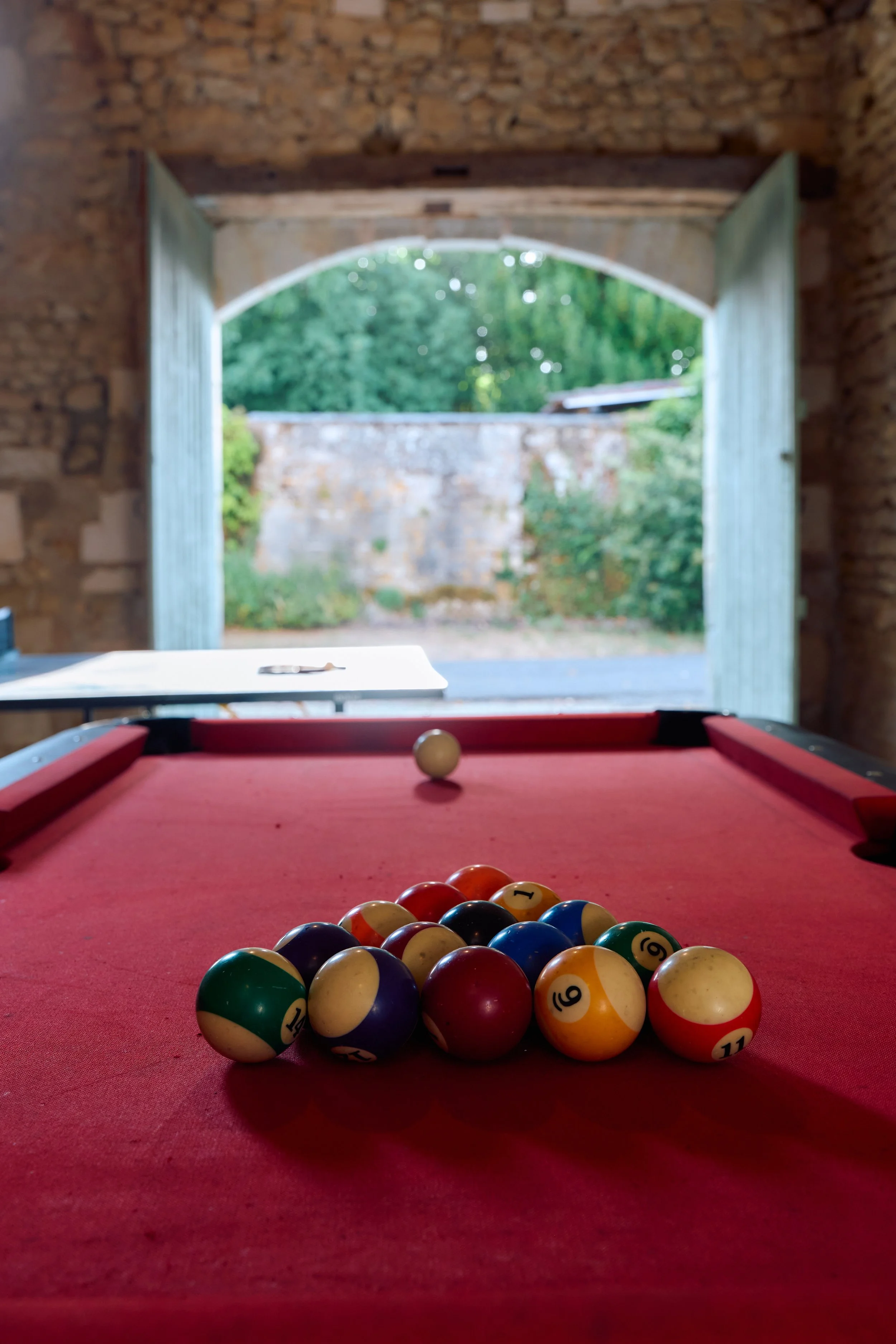 Pool table with billiard balls arranged on a red felt surface, viewed from the front, with a view outside through open double doors showing green trees and a stone wall.