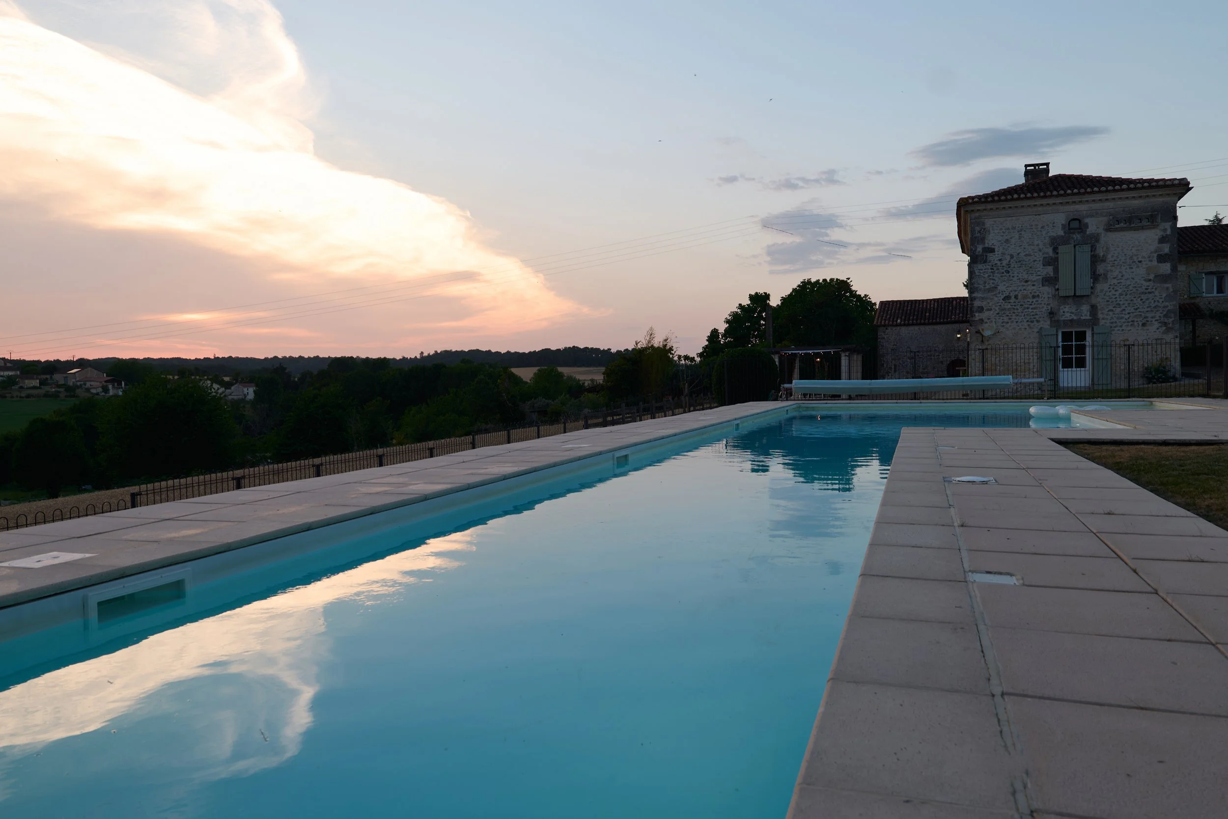 A serene outdoor swimming pool reflecting a colorful sunset sky with clouds, adjacent to a stone house with closed shutters and a tiled roof, surrounded by a black iron fence and greenery.