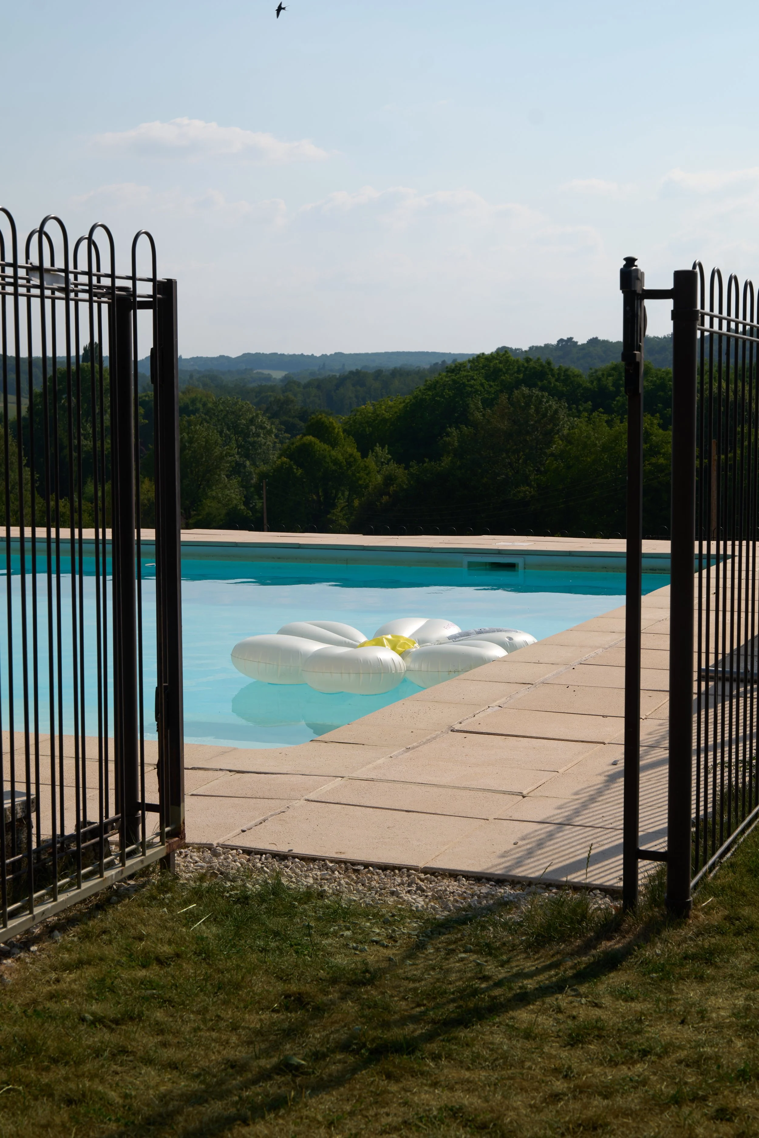 View of a swimming pool surrounded by a stone patio, with inflatable pool floats in the water. A black metal gate opens to the pool area, and rolling green hills with trees and a blue sky with some clouds are in the background.