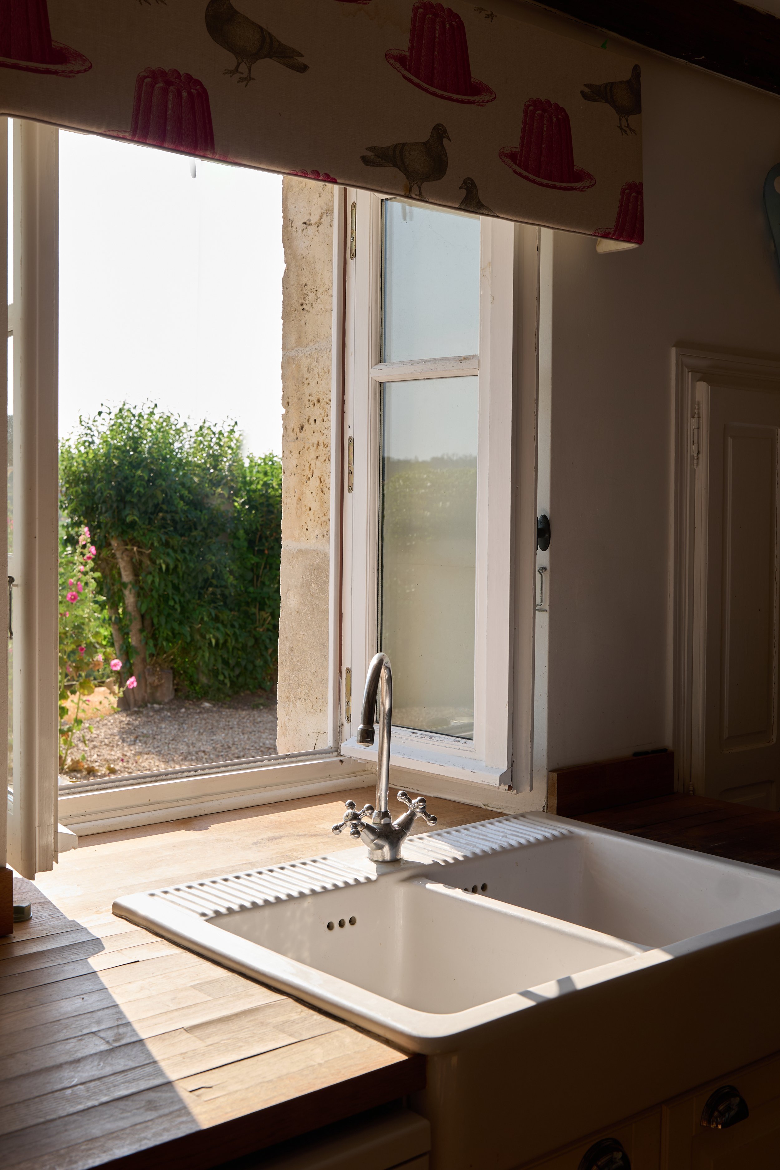 View of an open window in a kitchen with a white farmhouse sink and a silver faucet, overlooking a garden with greenery and flowers.
