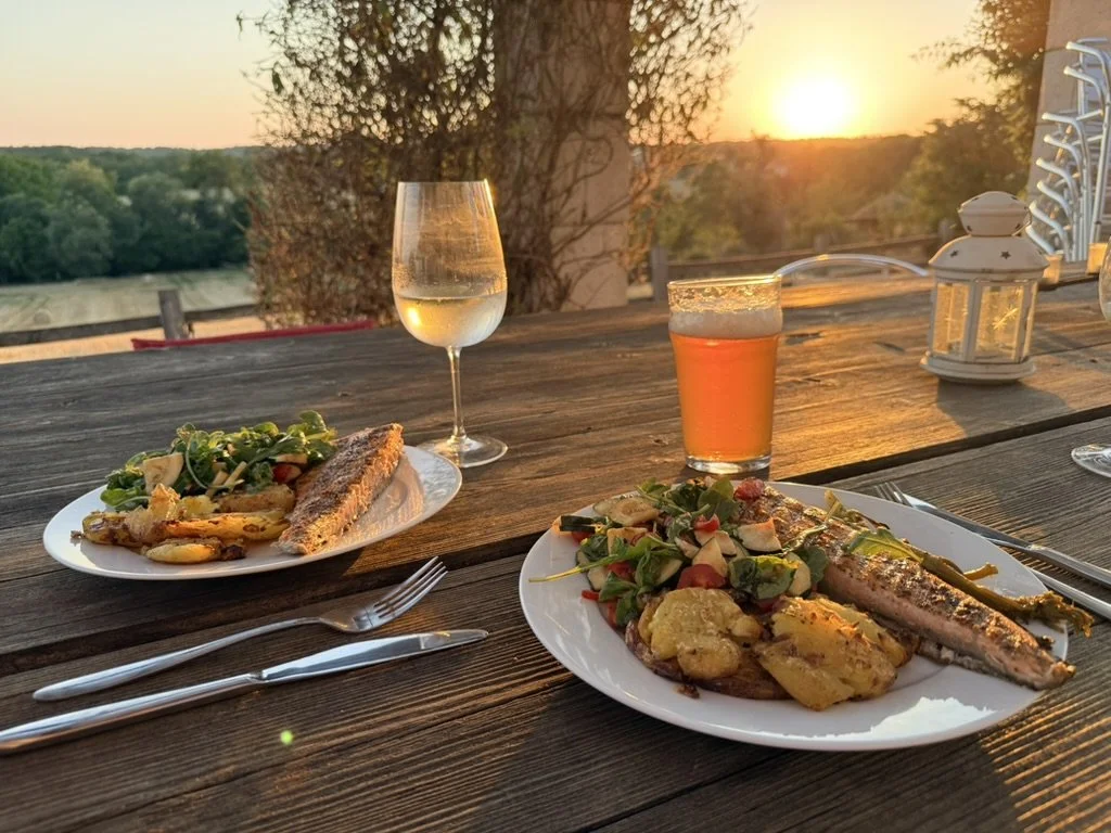 Outdoor dinner setting at sunset with plates of grilled fish, potatoes, and salad, a glass of white wine, a glass of beer, and lantern on a wooden table overlooking a natural landscape.