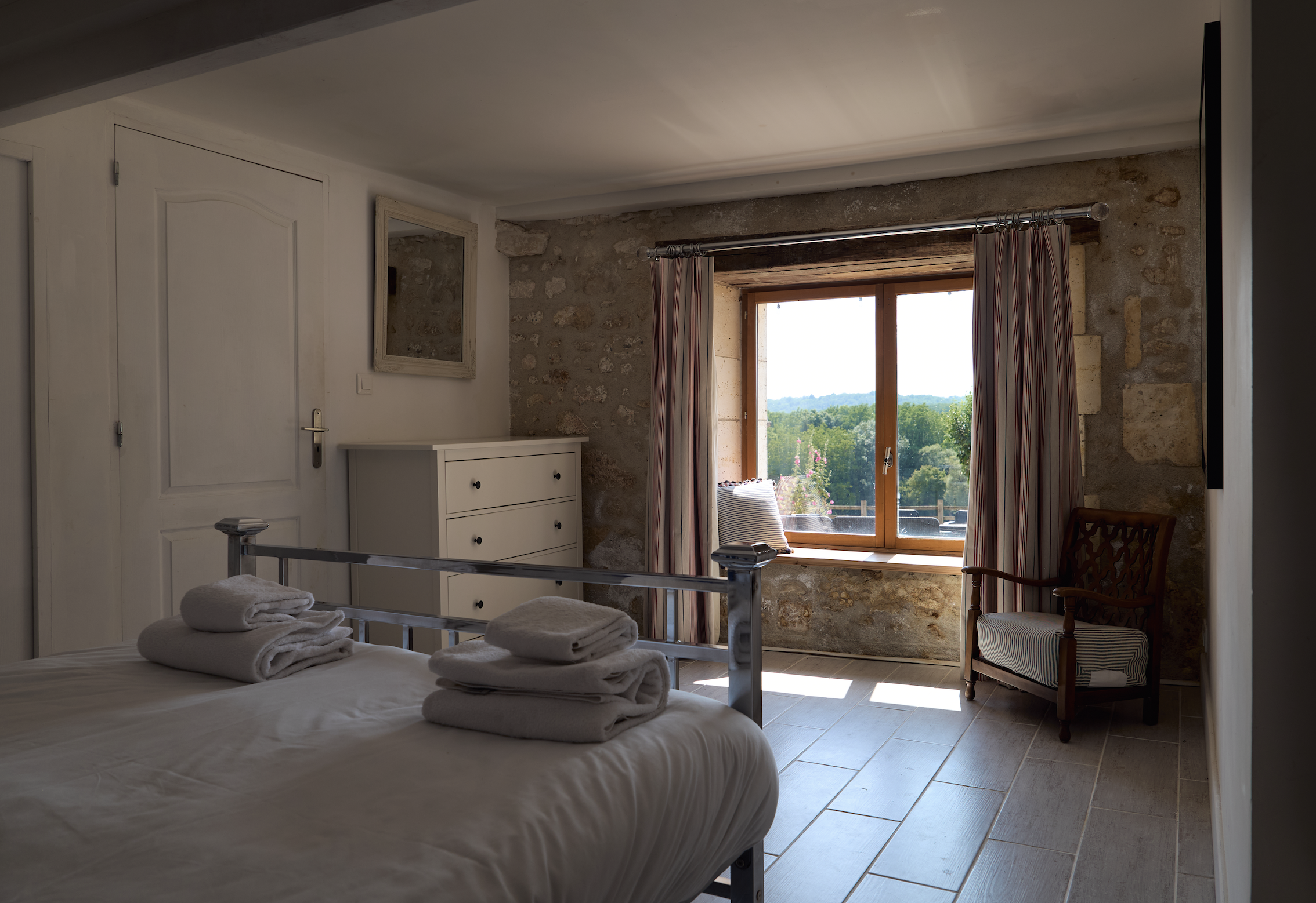 A cozy bedroom with a large window showing a scenic view of green trees and a clear sky. The window has striped curtains, and there is a white dresser and a wooden armchair with a cushion in the room. The bed has neatly folded towels on top.