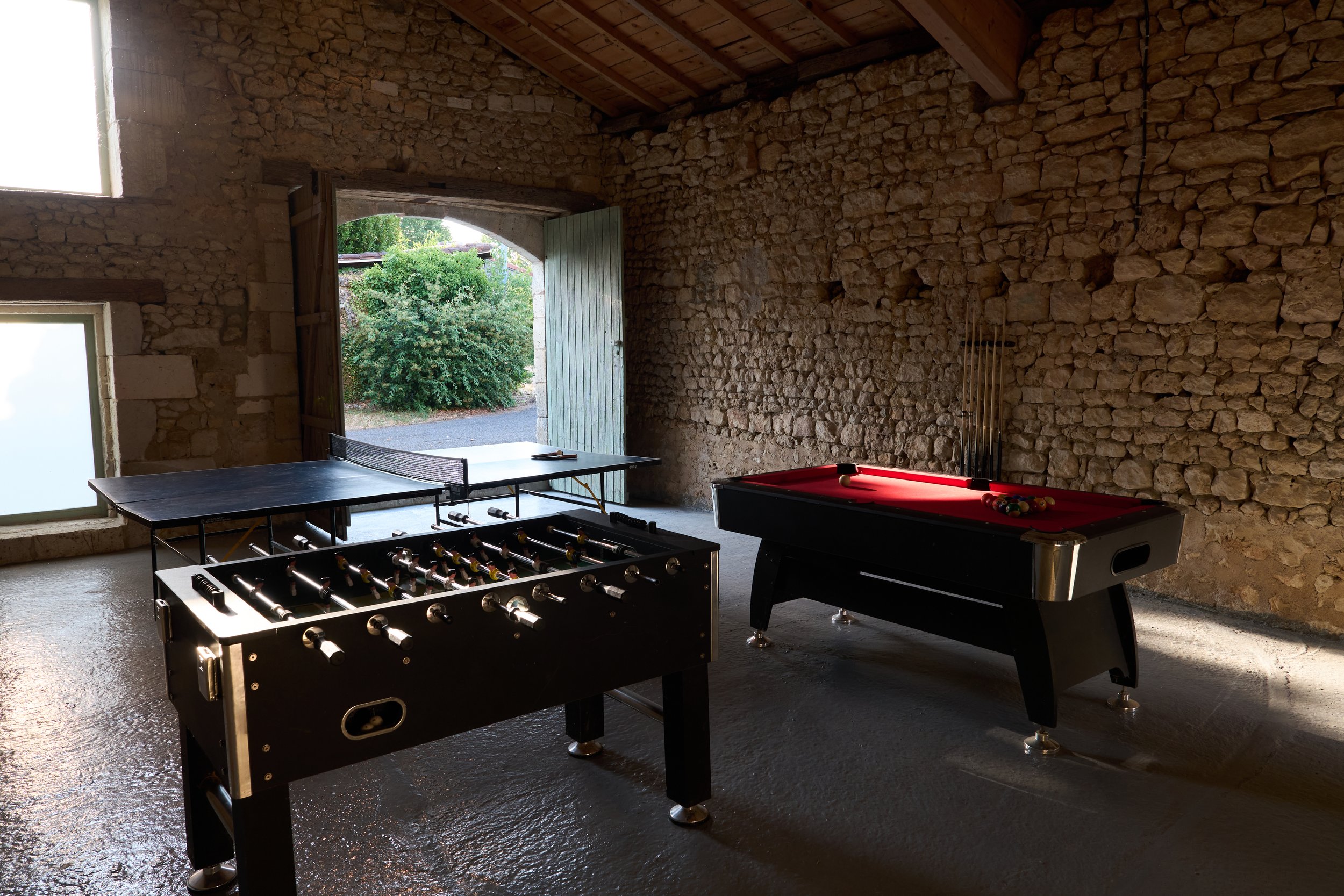Interior of a rustic stone barn with an open door leading outside, featuring a view of greenery. Inside, there is a foosball table, a pool table, a ping pong table, and a dartboard.