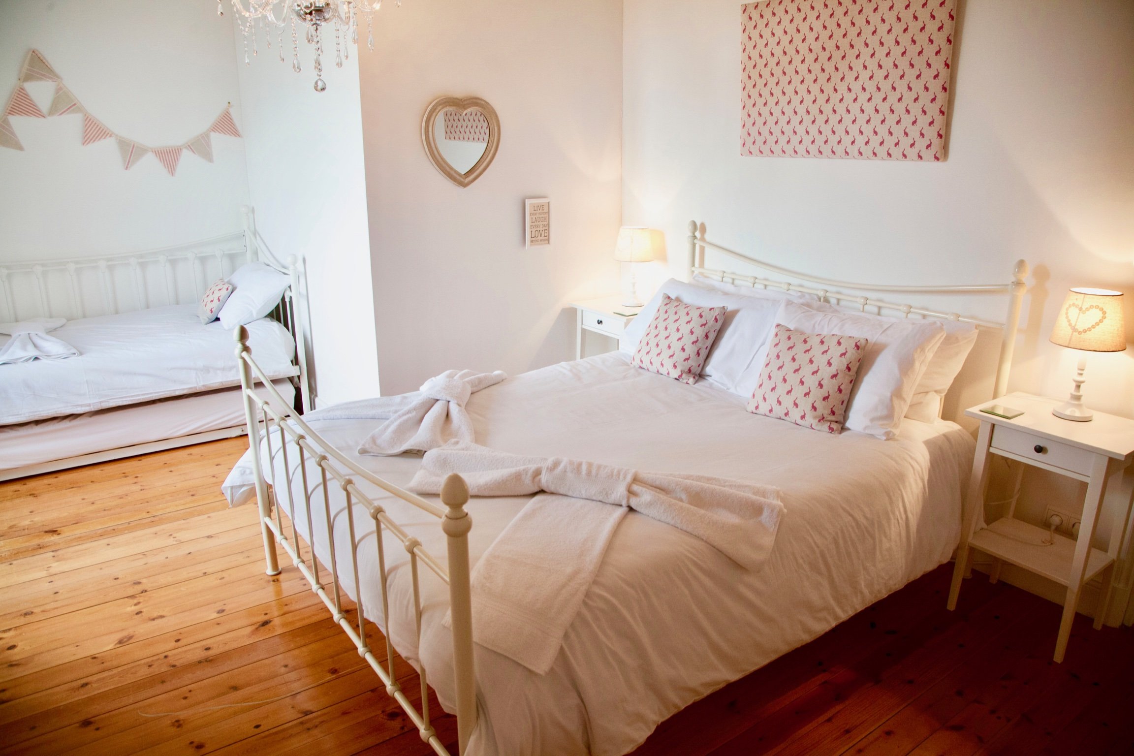A cozy bedroom with two white metal beds, decorated with matching pillows that have pink patterns. The room has wooden flooring, soft lighting from lamps, and decorative wall hangings, including a heart-shaped mirror, a framed quote, and pink banners.