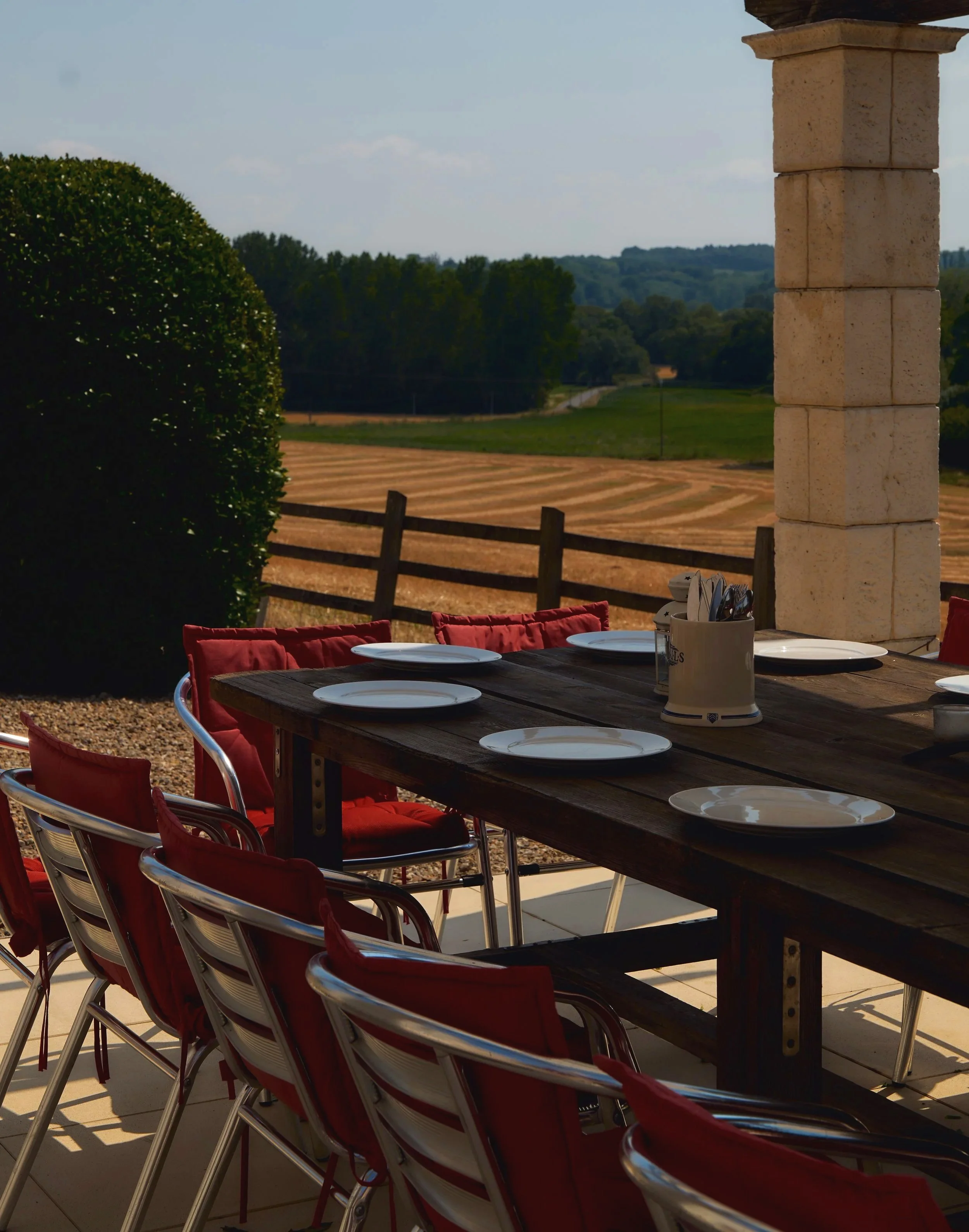 Outdoor patio table set with plates and utensils, overlooking a field and trees under a partly cloudy sky.