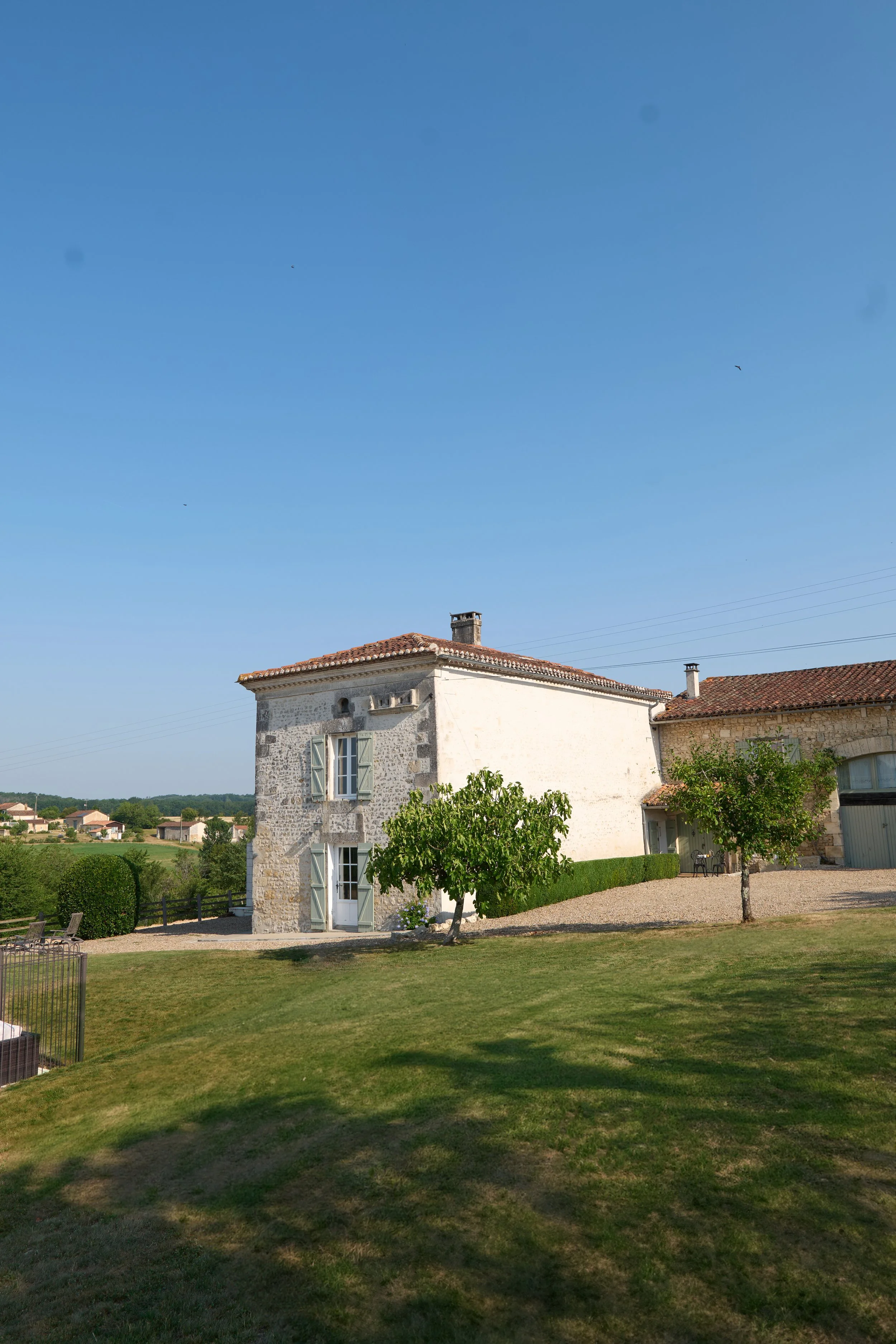 A white stone house with green shutters and a red tiled roof in a lush green yard with trees, bushes, benches, and a gravel pathway under a clear blue sky.