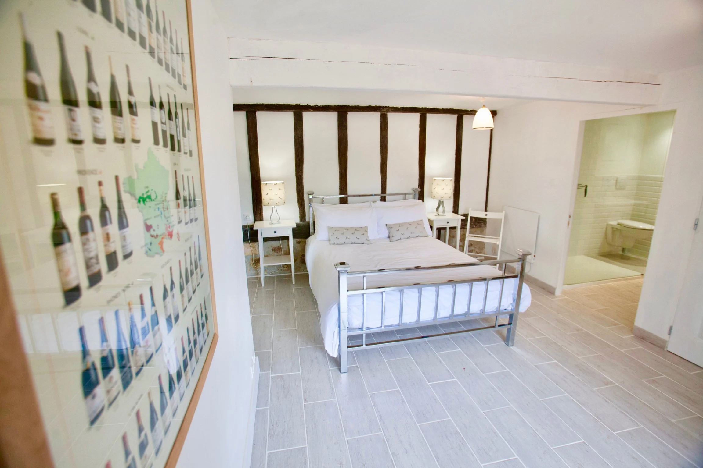 A bedroom with a white bed, nightstands with lamps, and a rustic wooden headboard. To the right, there is an open door leading to a bathroom with a white toilet and brick wall. To the left, a framed poster of wine bottles hangs on the wall.