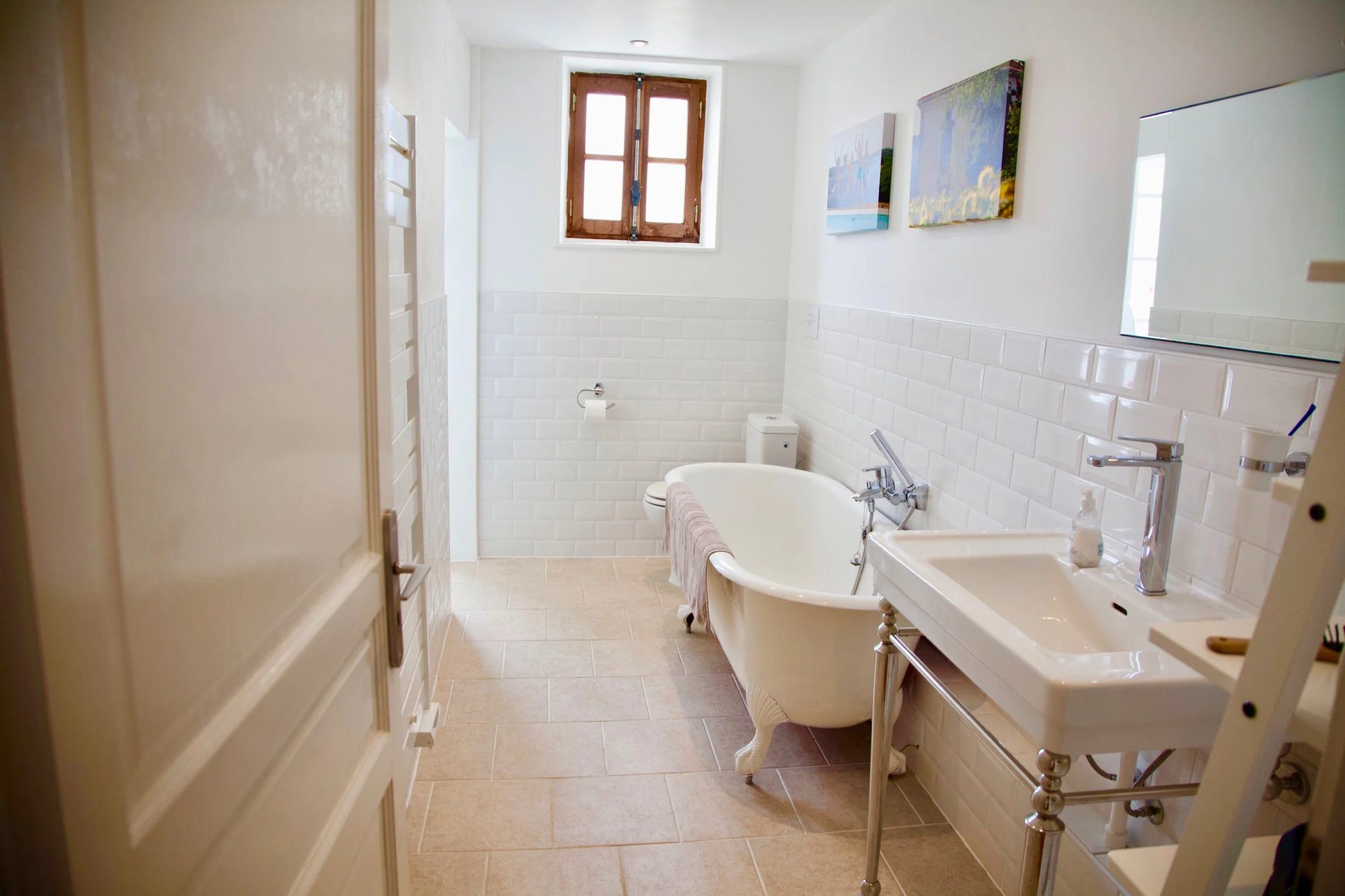 A small bathroom with a white bathtub, a toilet, and a sink. There is a window with wooden shutters, and two art paintings hang on the white wall. The floor has beige tiles.