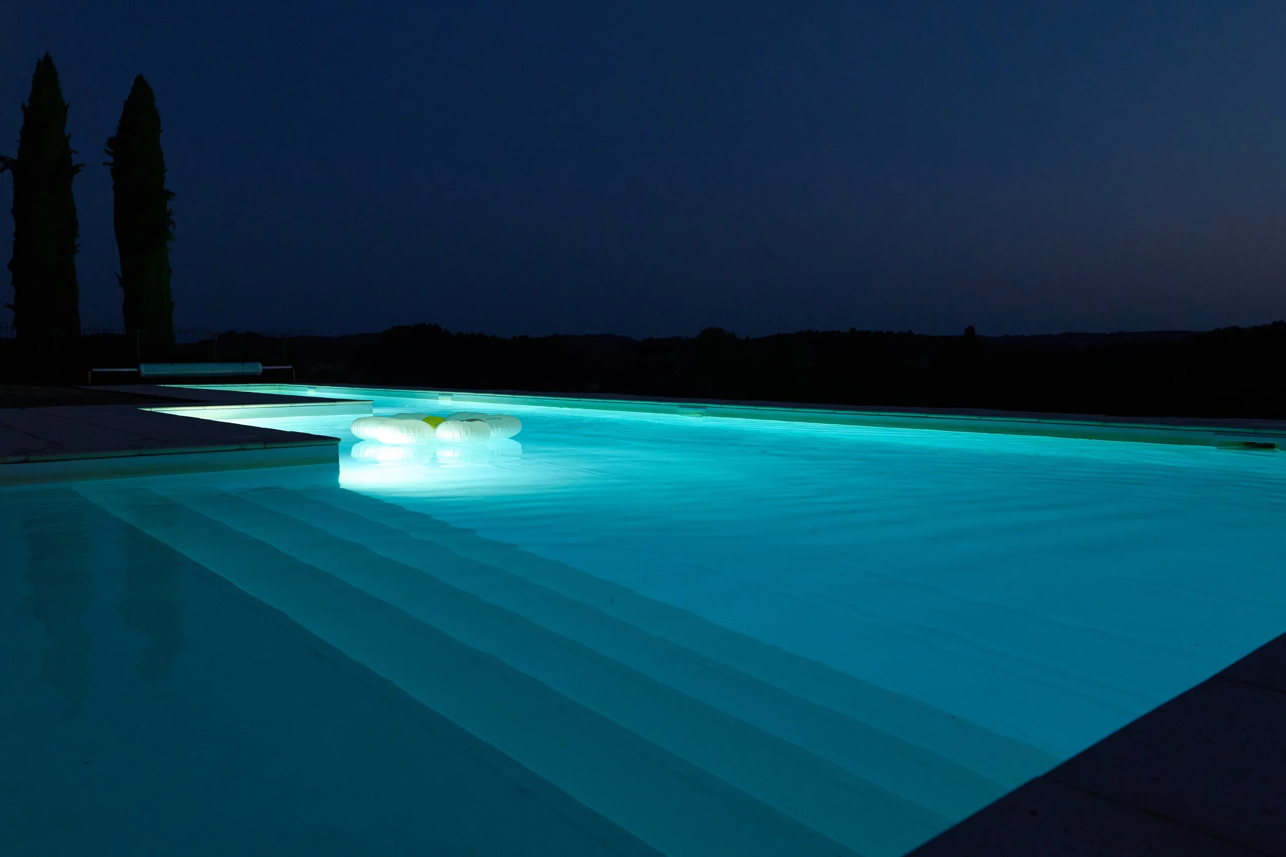 An illuminated swimming pool at night with a dark sky, two tall trees, and a hill in the background.