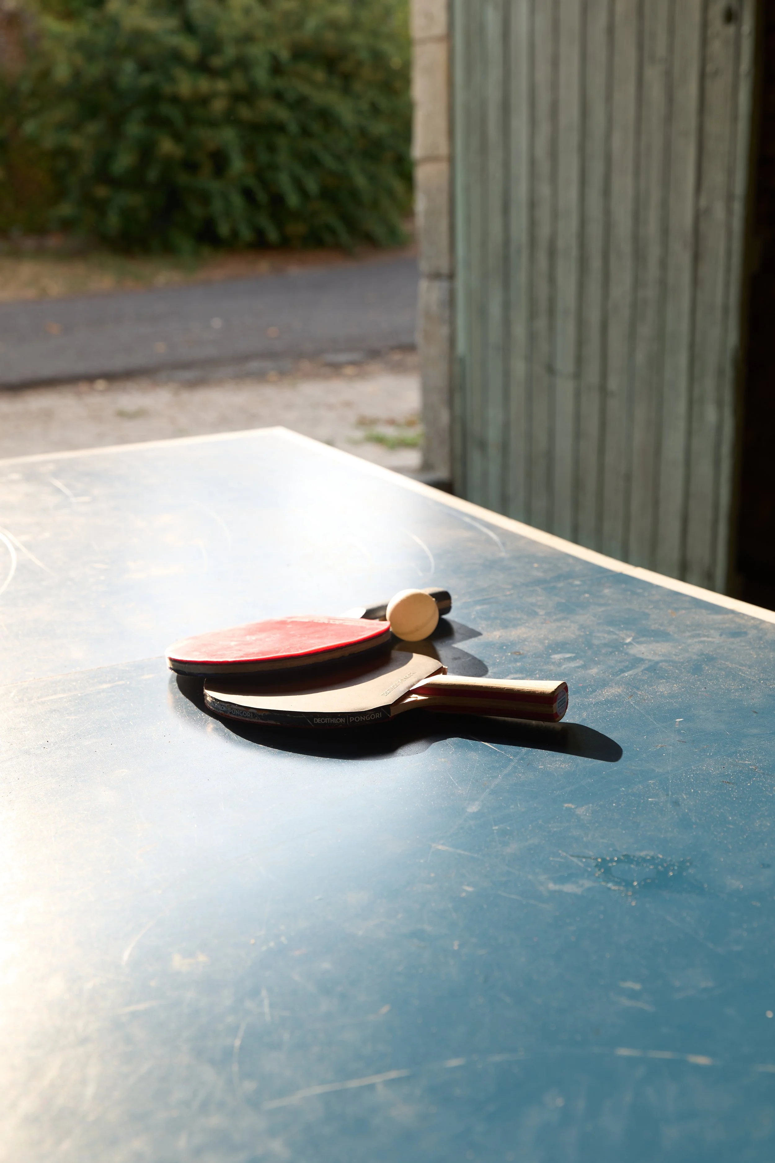 Table tennis paddles and ball on a weathered outdoor table, with a blurred background of hedges, pavement, and a wooden wall.