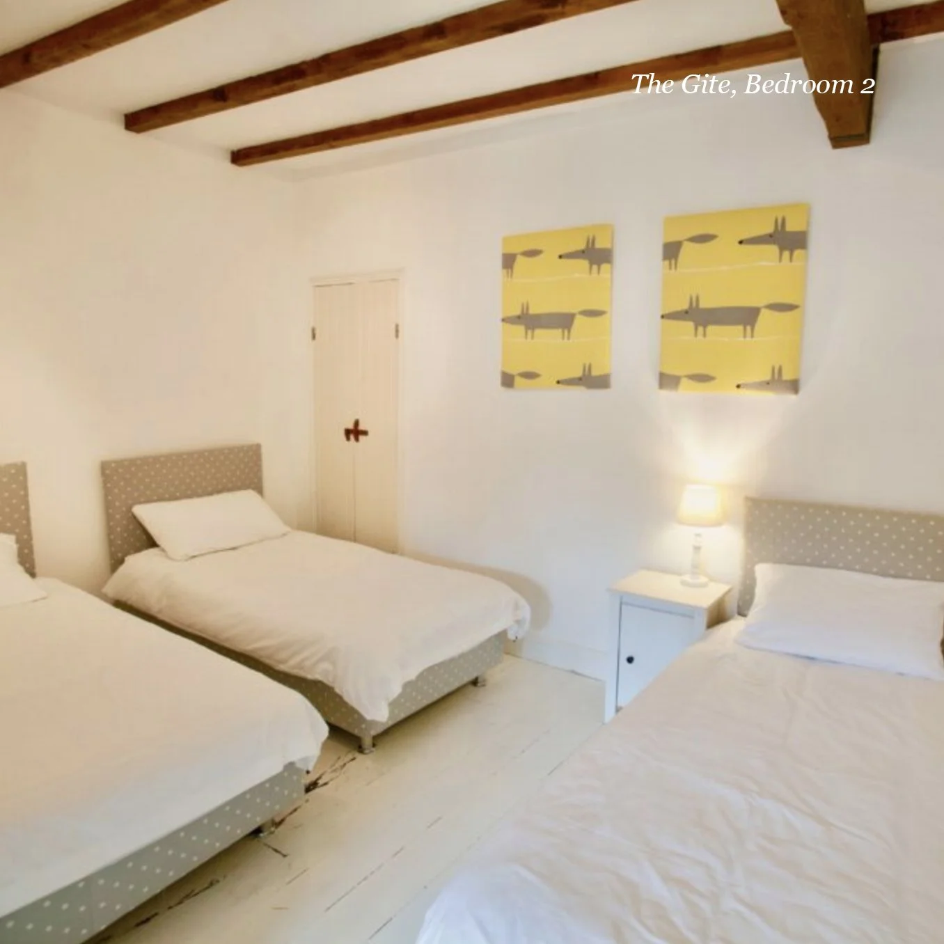 A bedroom with three single beds, white bedding, polka-dotted headboards, a white bedside table with a lamp, and wall art featuring stylized animal designs.