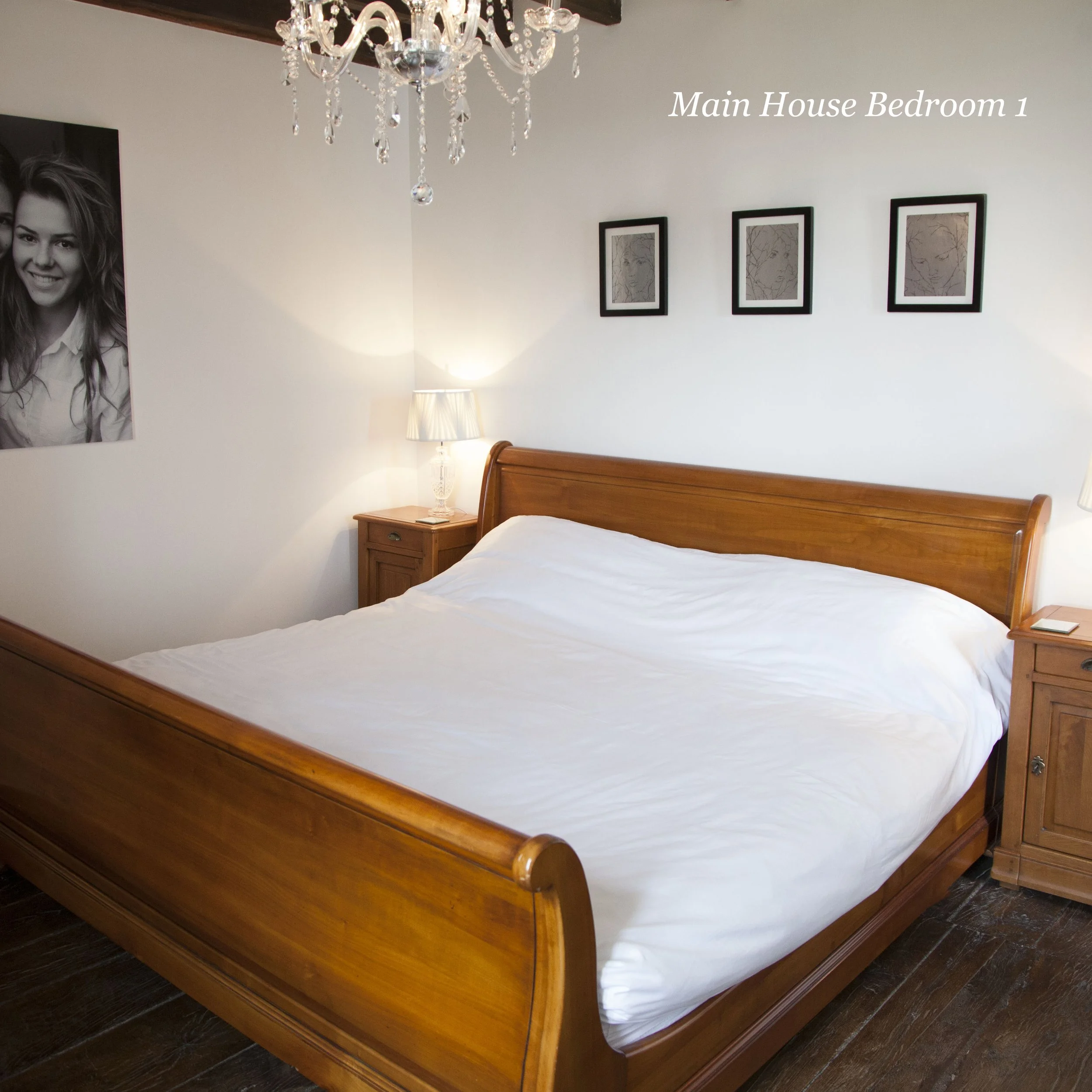 A wooden bed frame with a white bedspread, a wooden nightstand with a lamp, framed artwork on the wall, a chandelier hanging from the ceiling, and a large black-and-white portrait of a smiling woman on the wall.