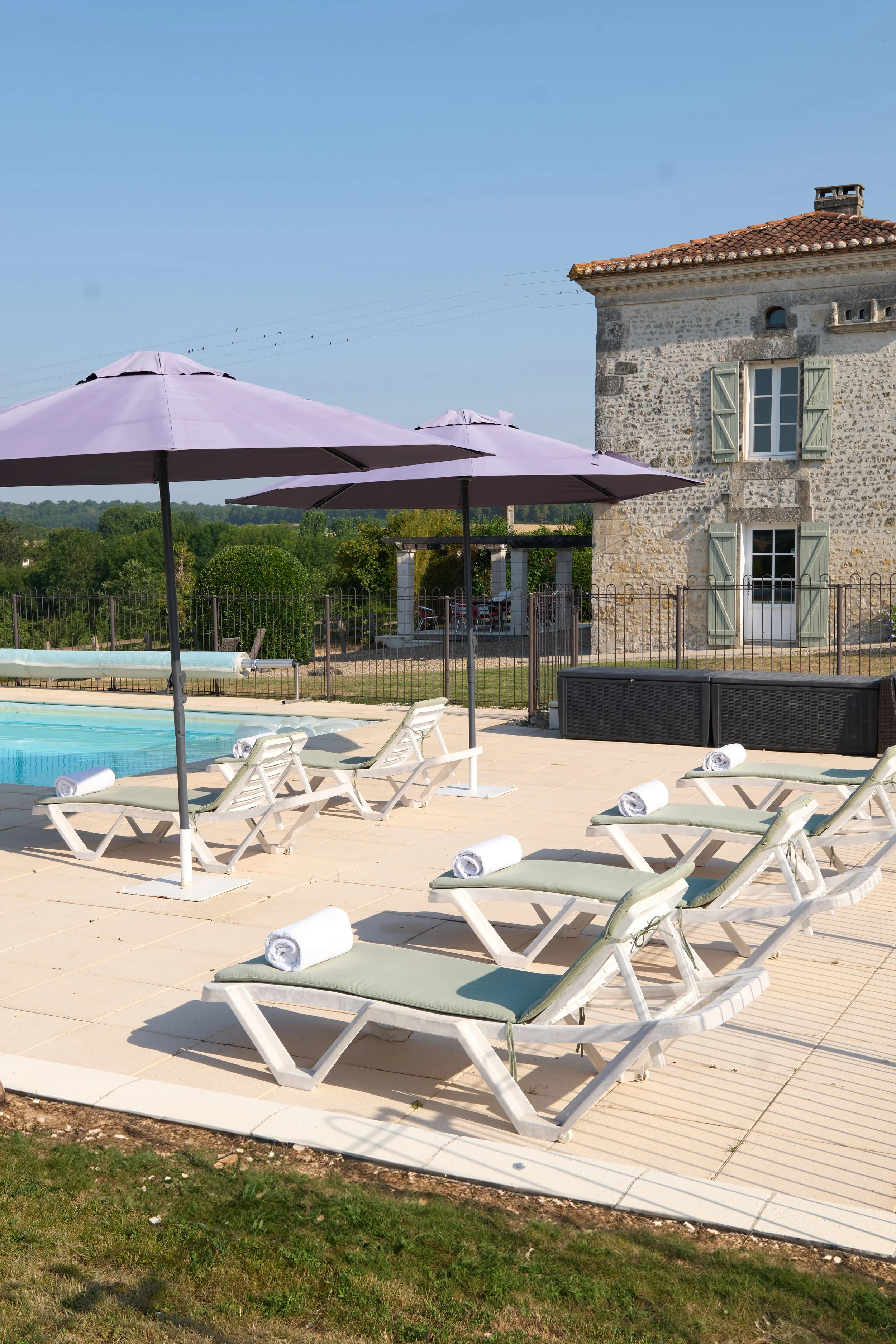 Swimming pool area with several white lounge chairs and purple umbrellas, adjacent to a stone house with boarding shutters, in a lush green landscape under a clear blue sky.