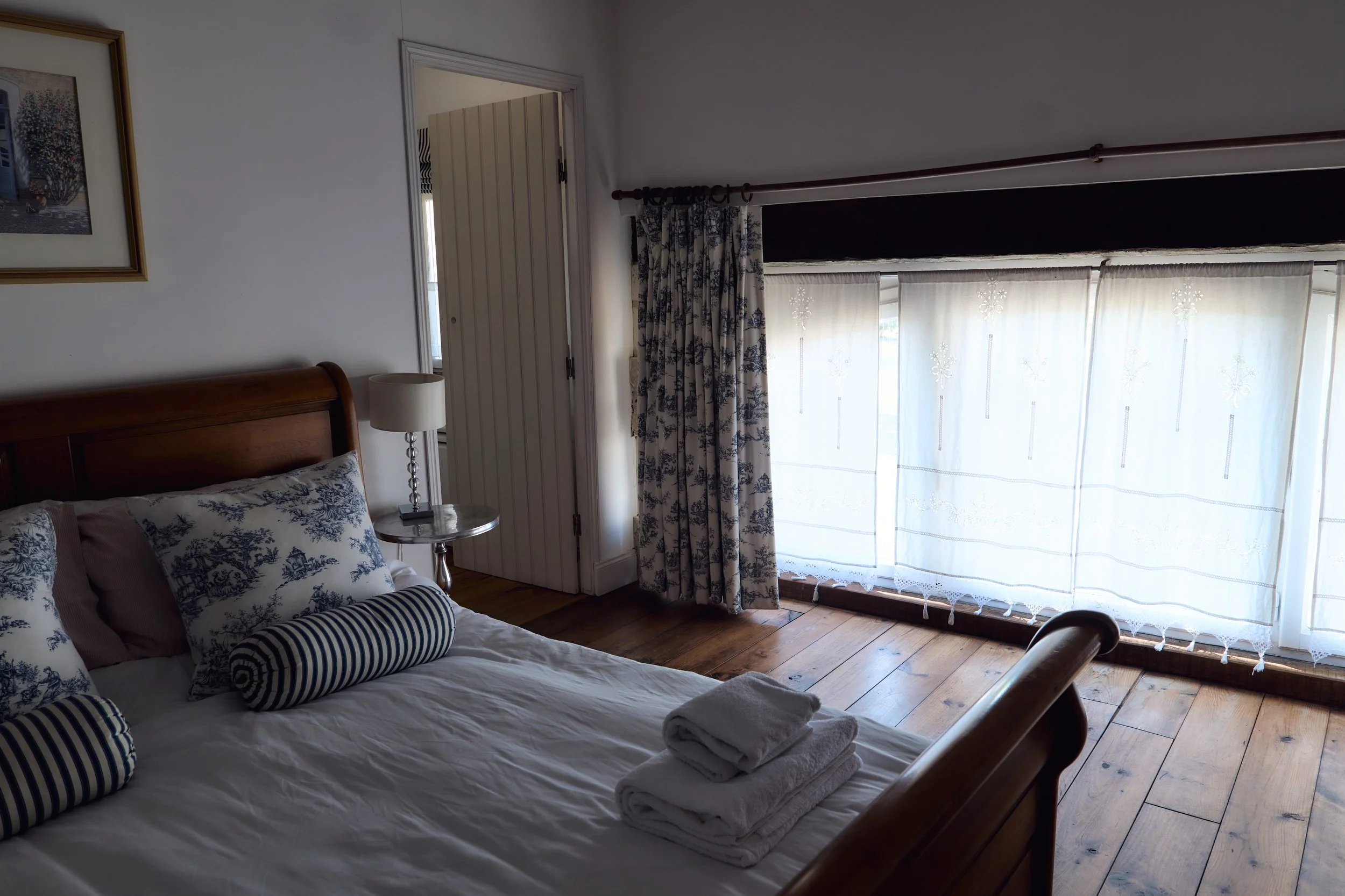 A bedroom with a wooden bed frame, patterned pillows, and two rolled towels on the bed. There is a nightstand with a white lamp, white curtains with a floral pattern, and sheer white drapes covering a large window.